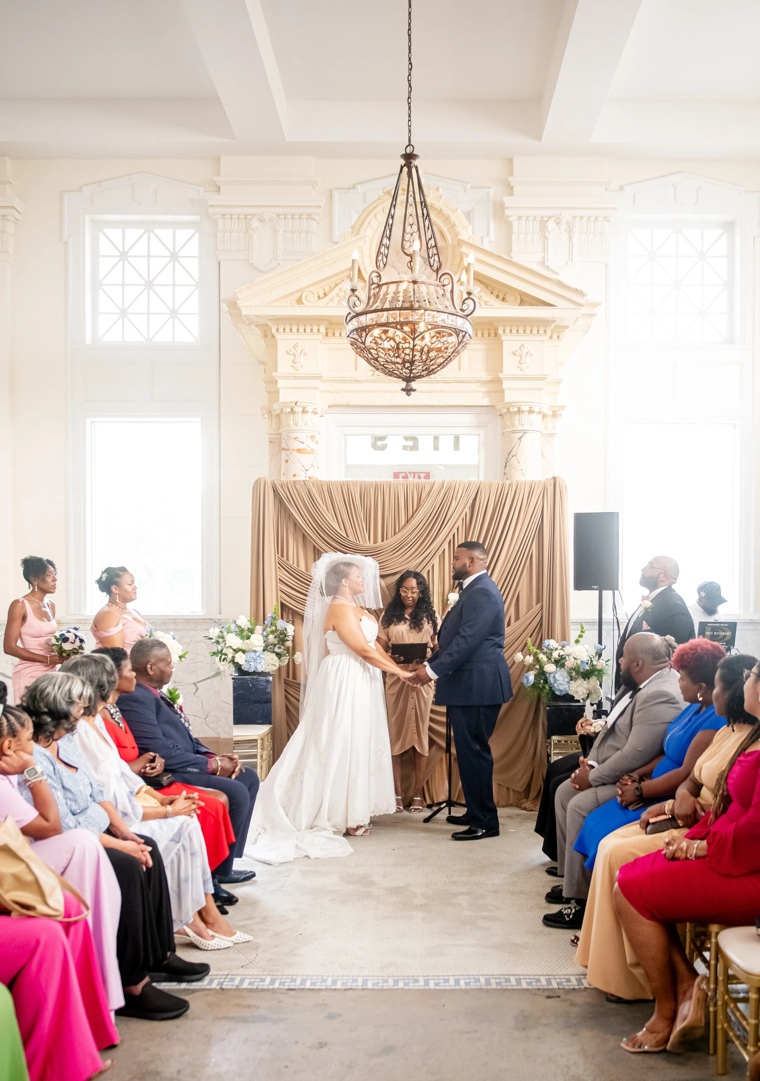 A wedding ceremony with a bride and groom exchanging vows in a bright, elegant room with high ceilings, large windows, and a decorative mantel. The bride is dressed in a white gown with a veil, and the groom is in a navy suit. Guests are seated on bo