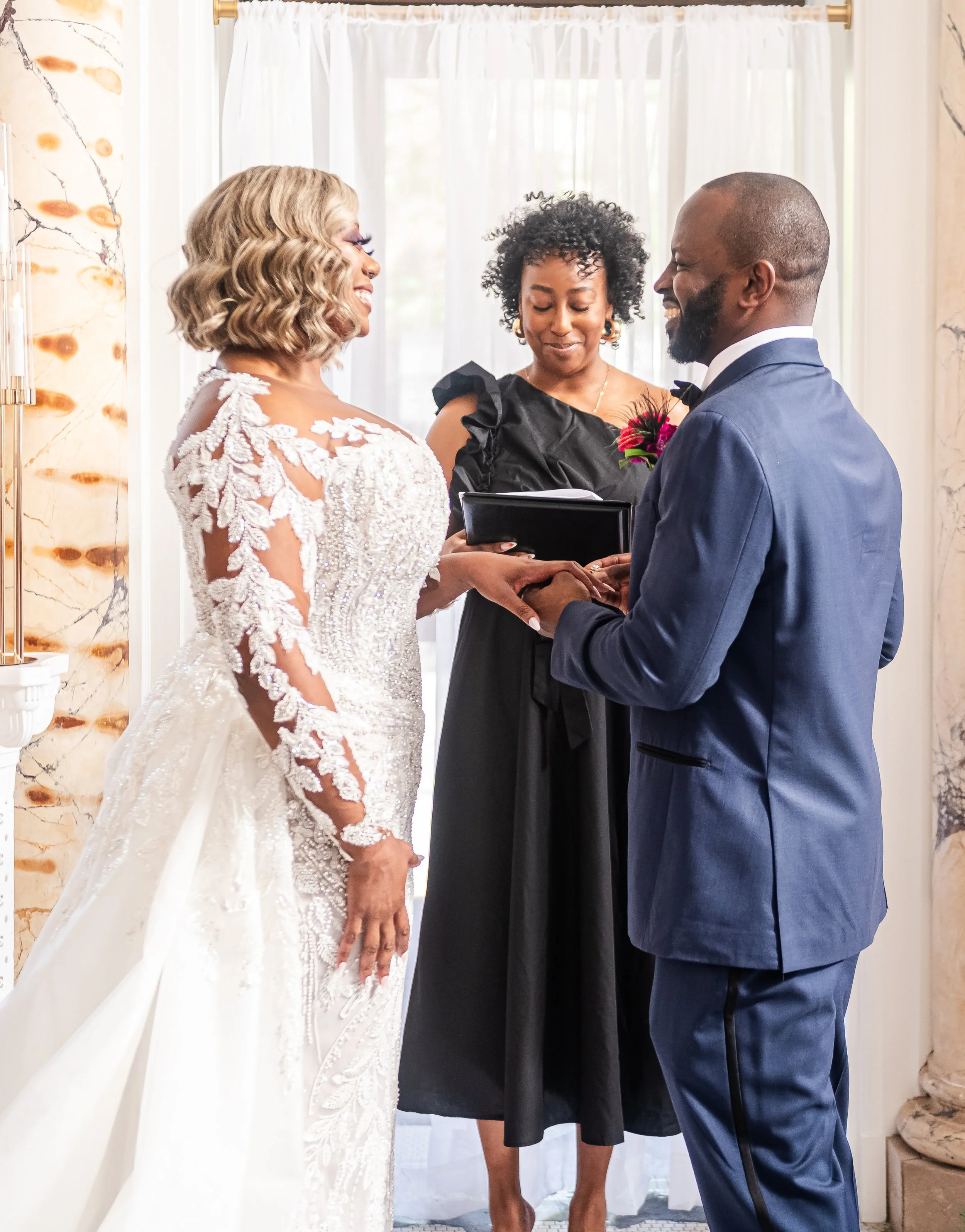 A couple getting married, holding hands and exchanging vows, with an officiant in the middle, set in a bright room with curtains and marble walls.
