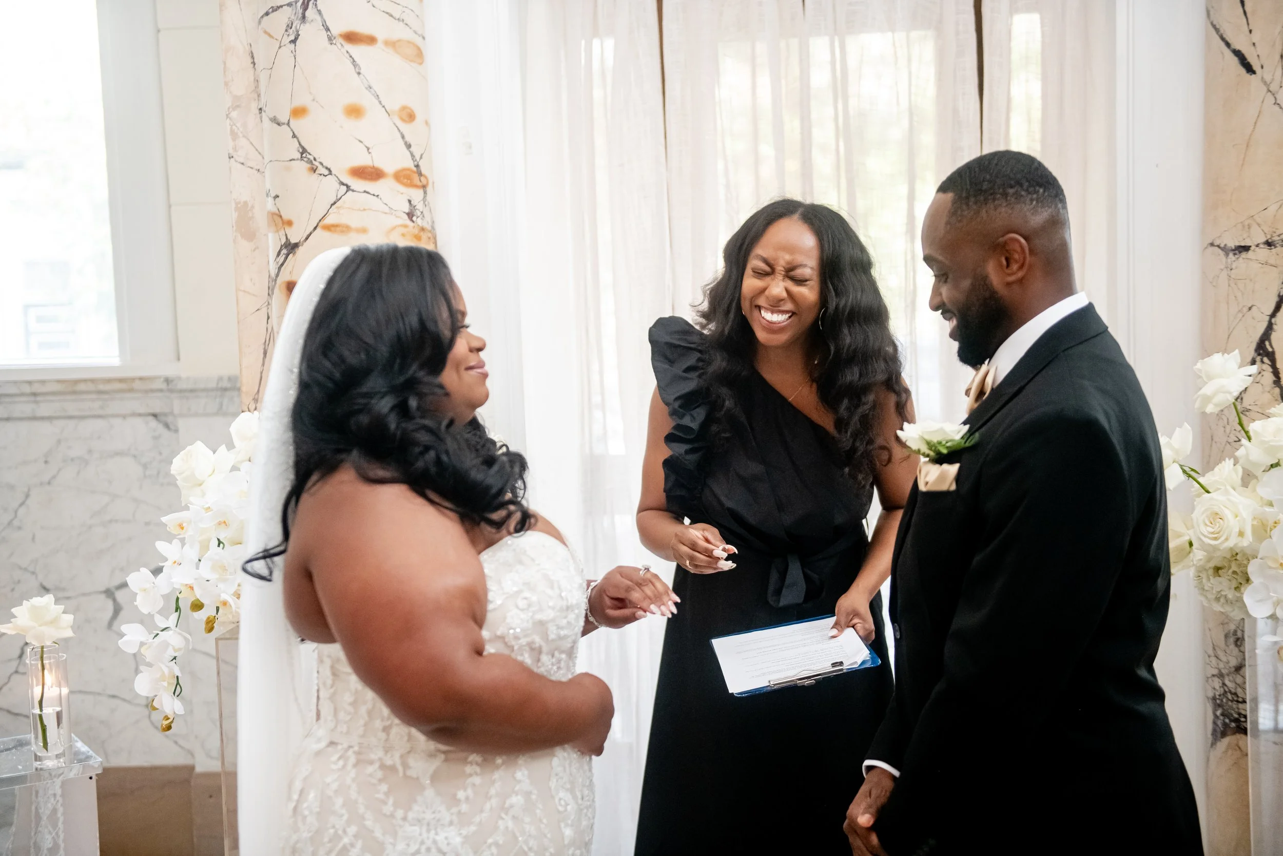 A bride and groom exchanging vows during a wedding ceremony with a officiant smiling between them. The bride is wearing a white lace wedding dress, and the groom is dressed in a black suit with a white shirt and beige bow tie. They are indoors with c