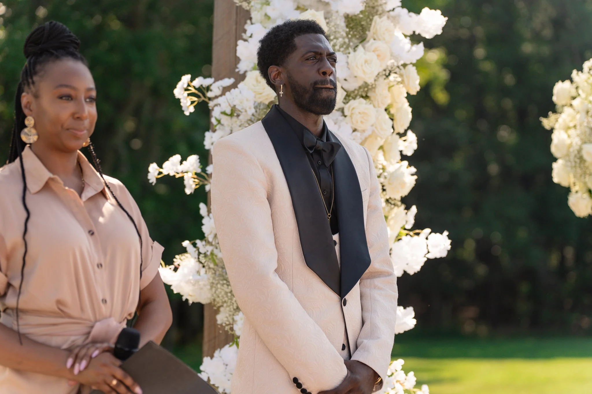 A man dressed in a white tuxedo with black lapel and bow tie, standing in front of a floral backdrop with white roses, during an outdoor wedding ceremony. A woman holding a microphone and clipboard is beside him.