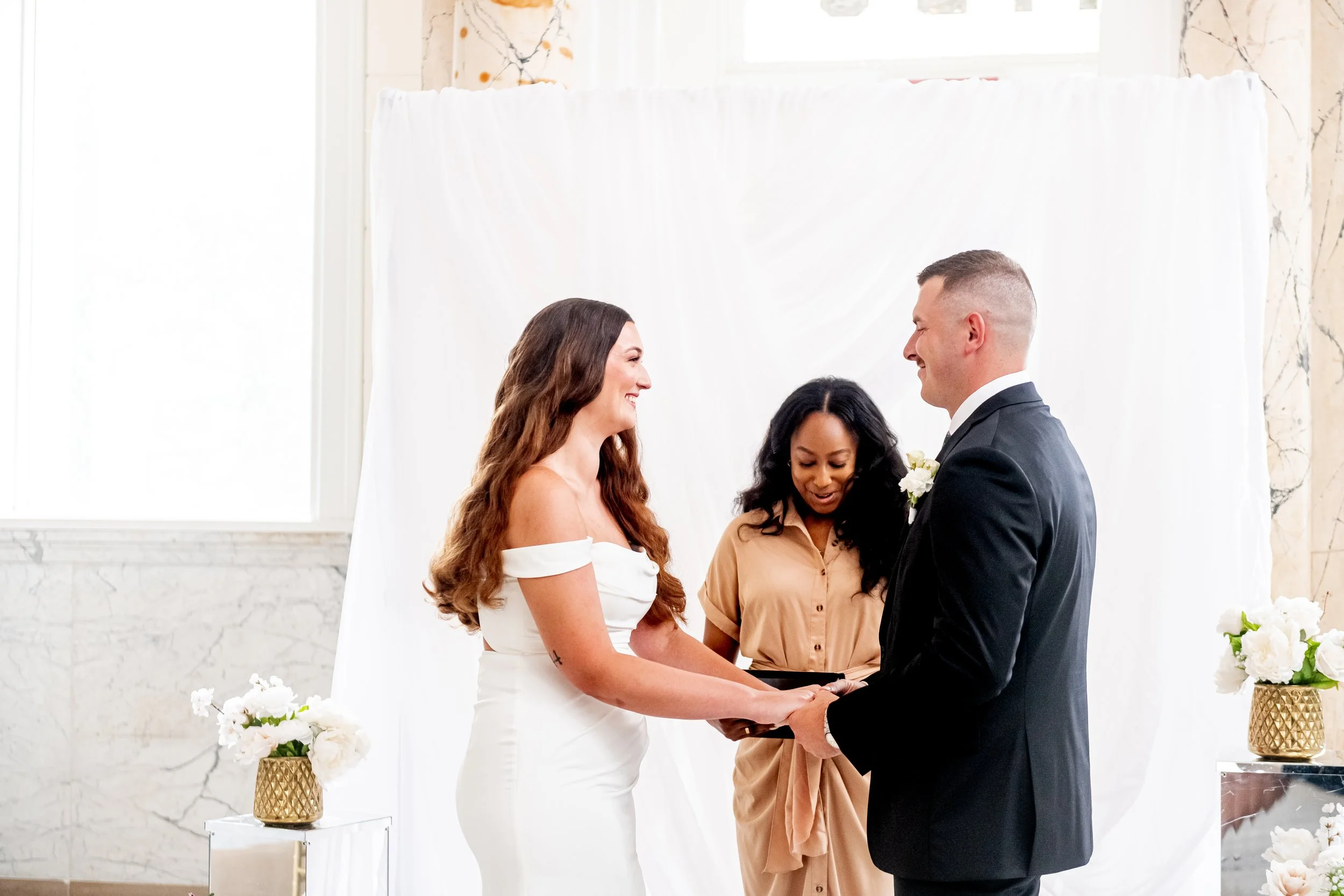 A couple getting married, exchanging vows, with a woman officiant standing between them, all smiling, inside a decorated venue with white drapes and floral arrangements.