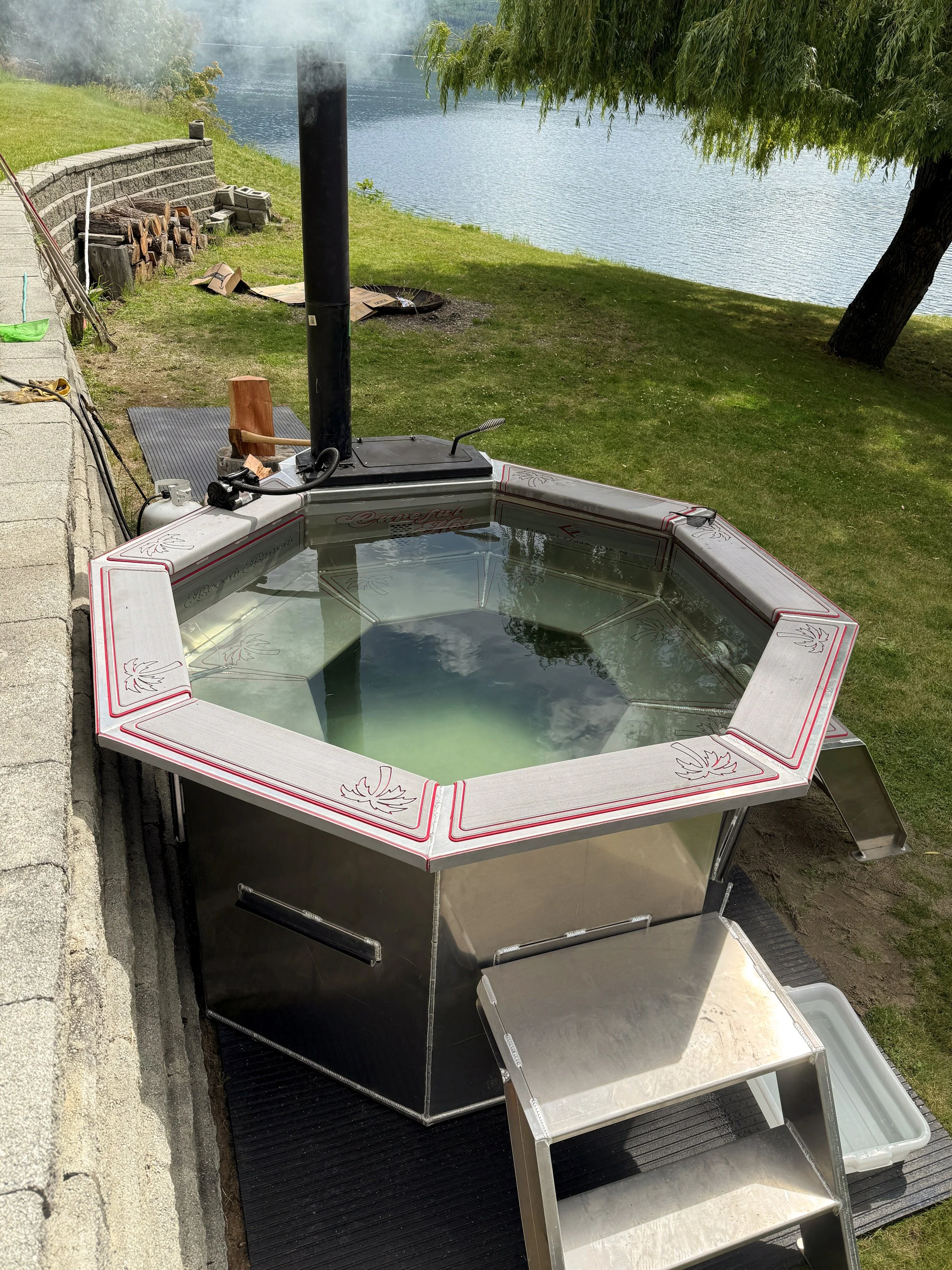 A metal outdoor hot tub filled with water, positioned on a black mat outside near a grassy area and a lake, with a small metal staircase leading into it and a wood-burning stove chimney pipe attached.