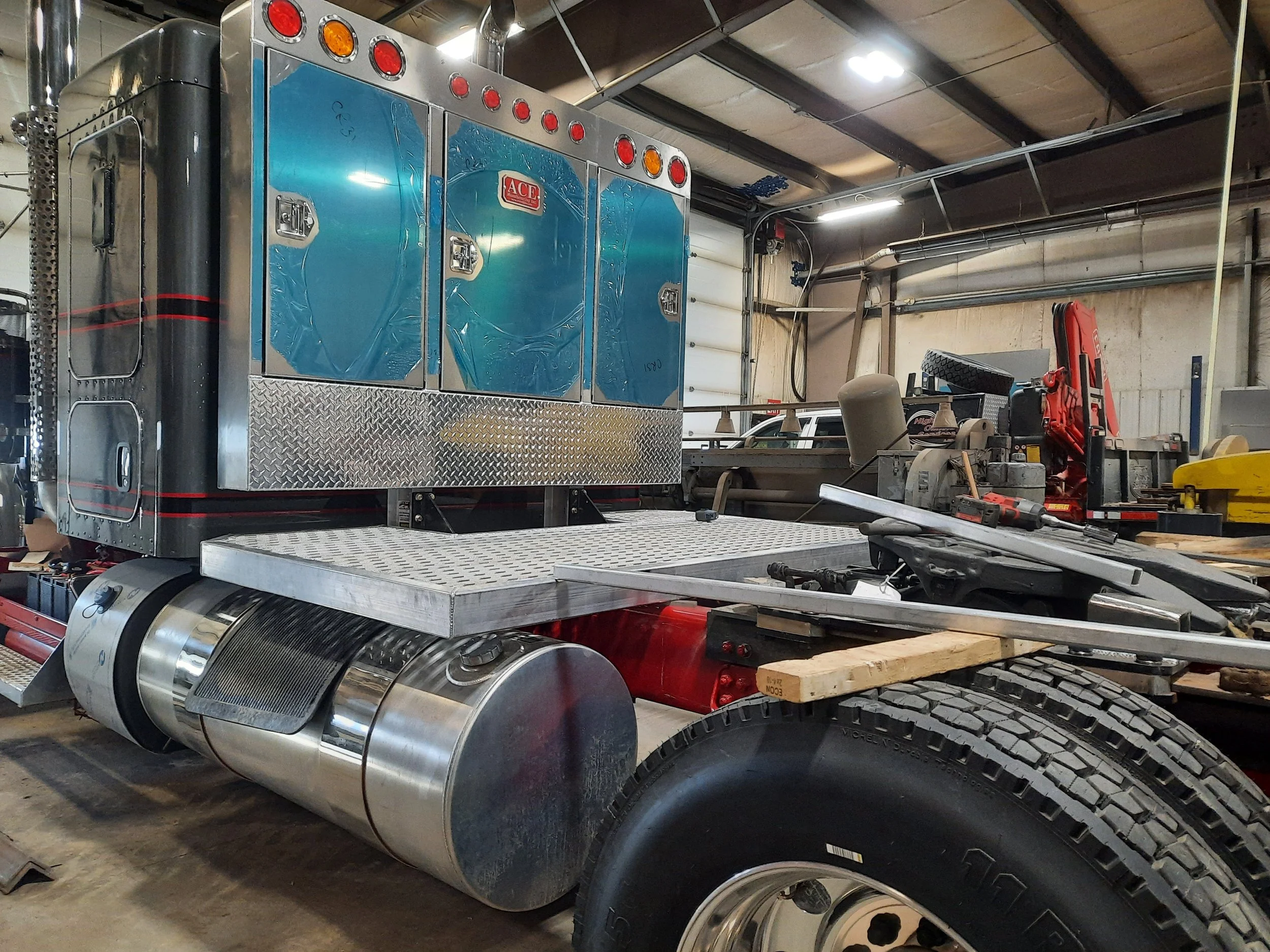 Close-up of a semi-truck's chassis showing a large chrome fuel tank, a metal platform, and a section of the rear wheel. The truck is inside a workshop with various tools and equipment visible.