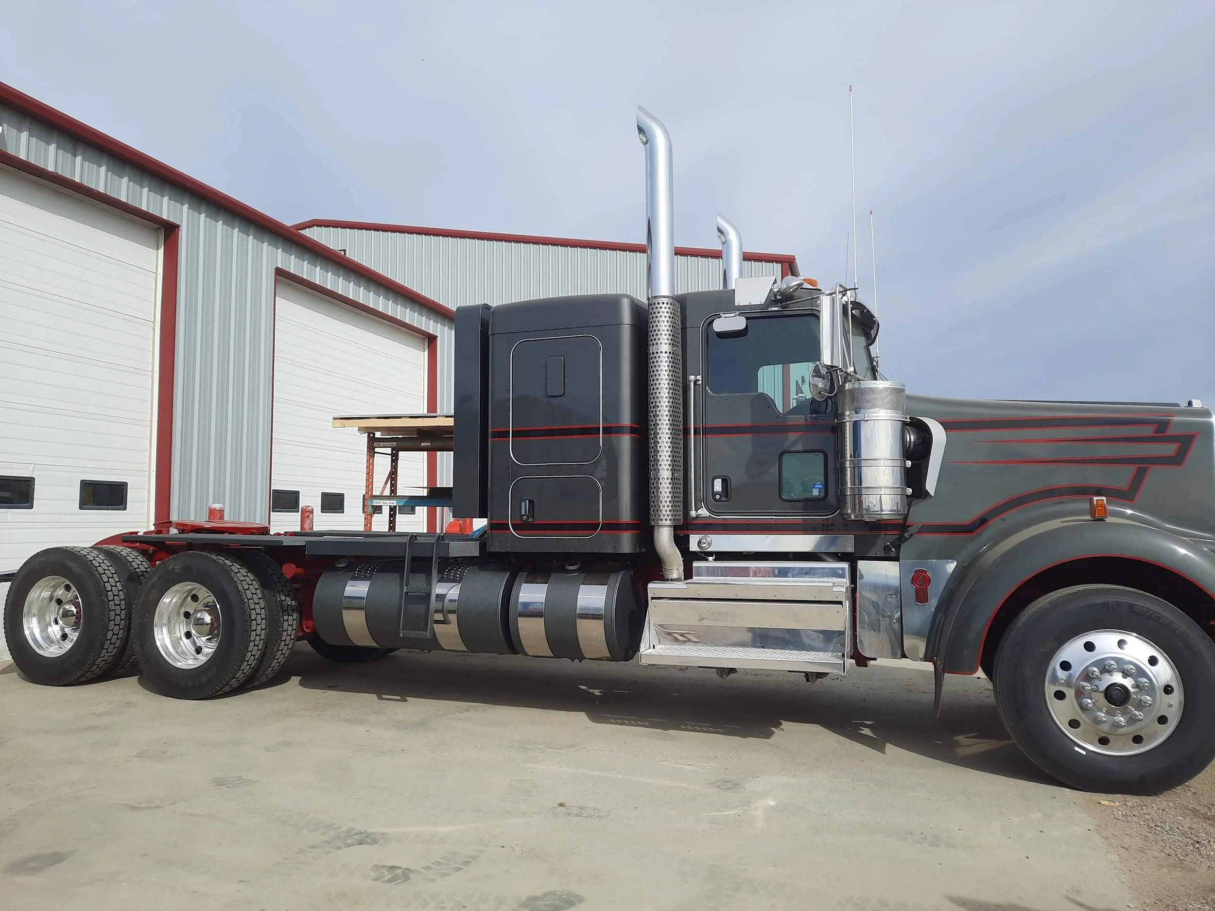 A semi-truck with a black and red design parked outside a large industrial garage. The truck has multiple rear tires, chrome exhaust stacks, and a metallic fuel tank.