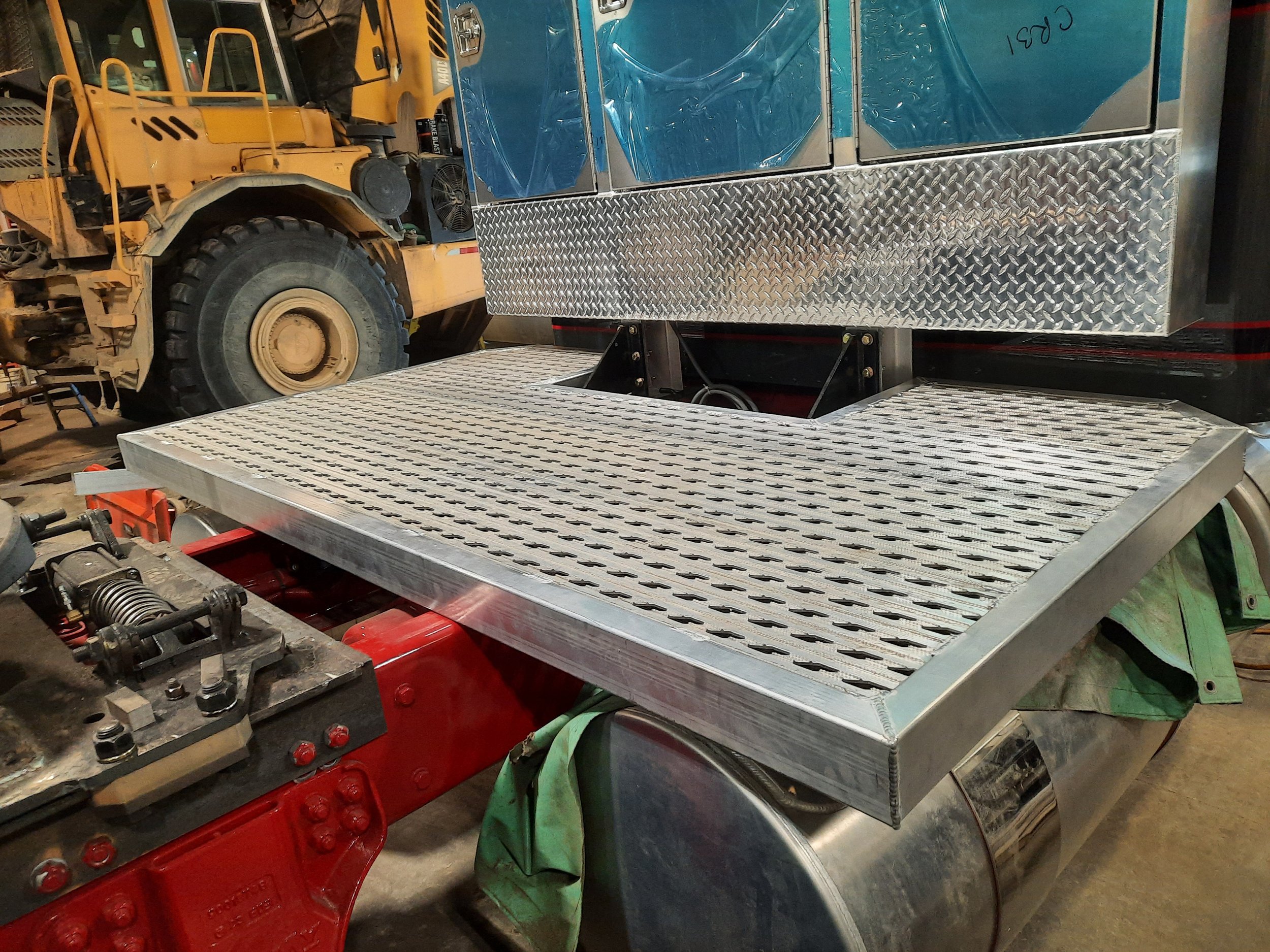 Close-up view of a metallic platform or step with a diamond plate pattern, attached to part of a vehicle or machinery, with a red component underneath and a large yellow construction vehicle in the background.