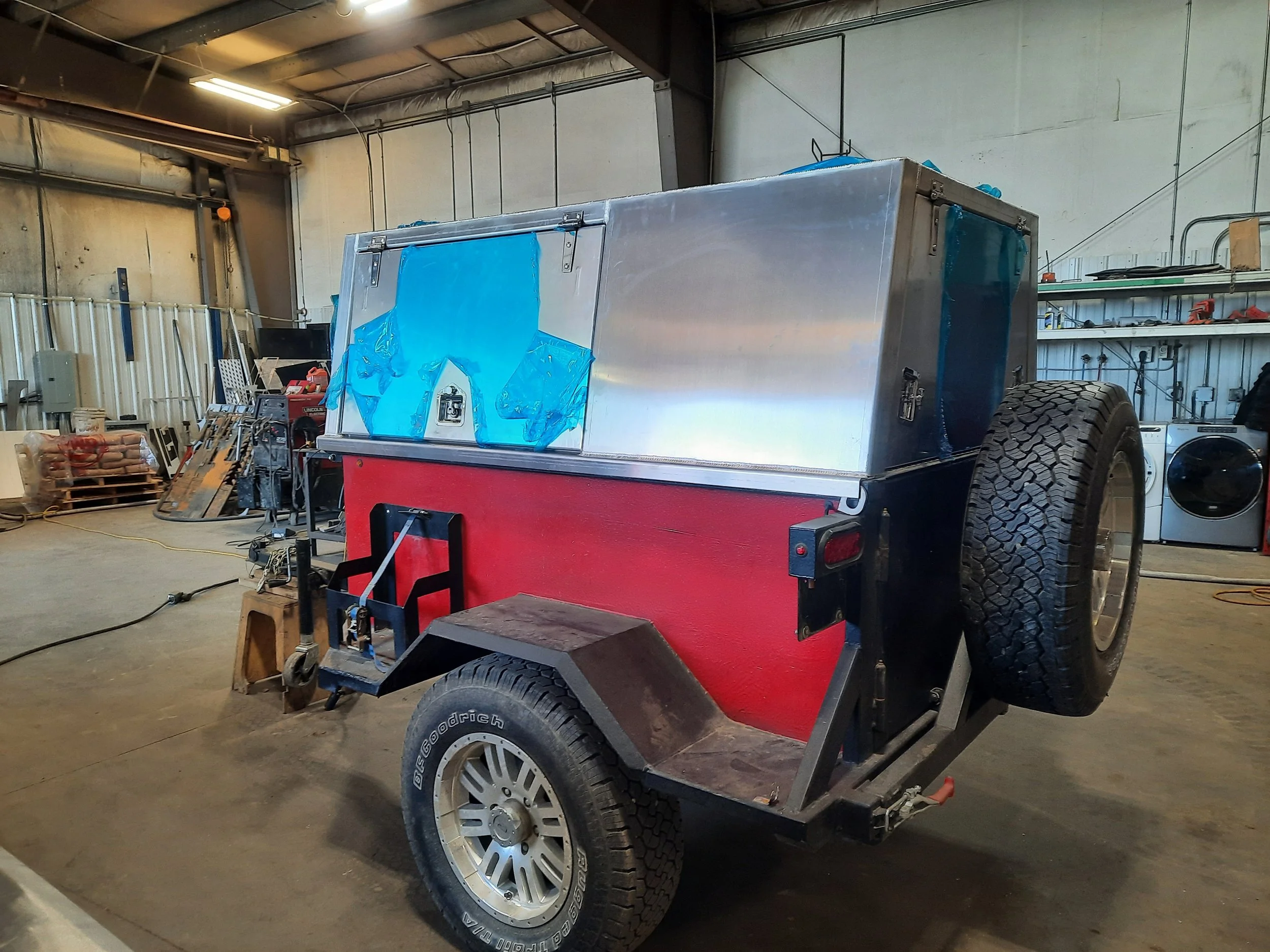 A small trailer with a red lower section and a silver upper section, parked inside a workshop. The trailer has large tires, doors on the front and side, and various tools and equipment are visible in the background.