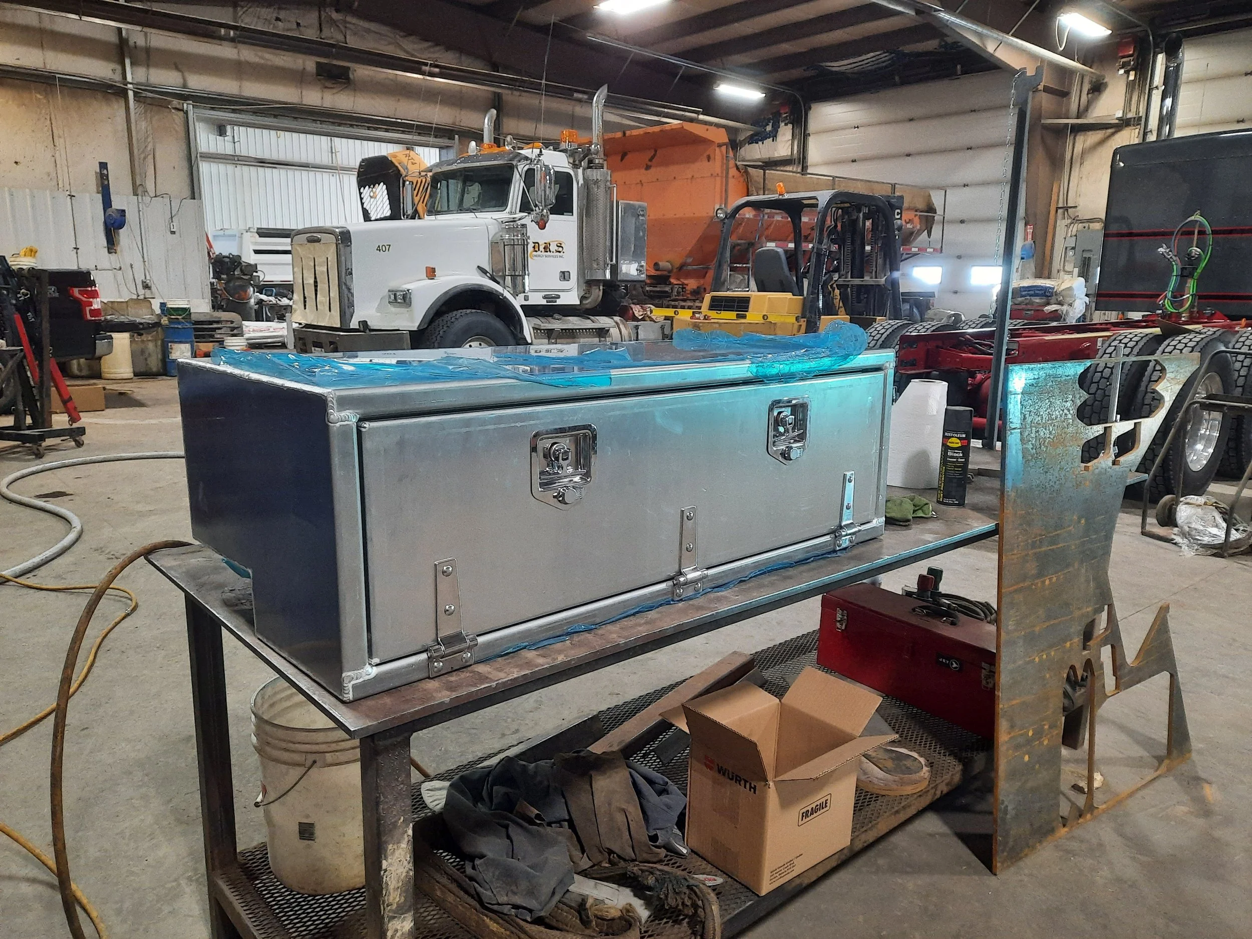 A workshop with a metal toolbox on a workbench, surrounded by tools, boxes, and cleaning supplies. In the background, large trucks and construction equipment are visible inside a garage or industrial building.