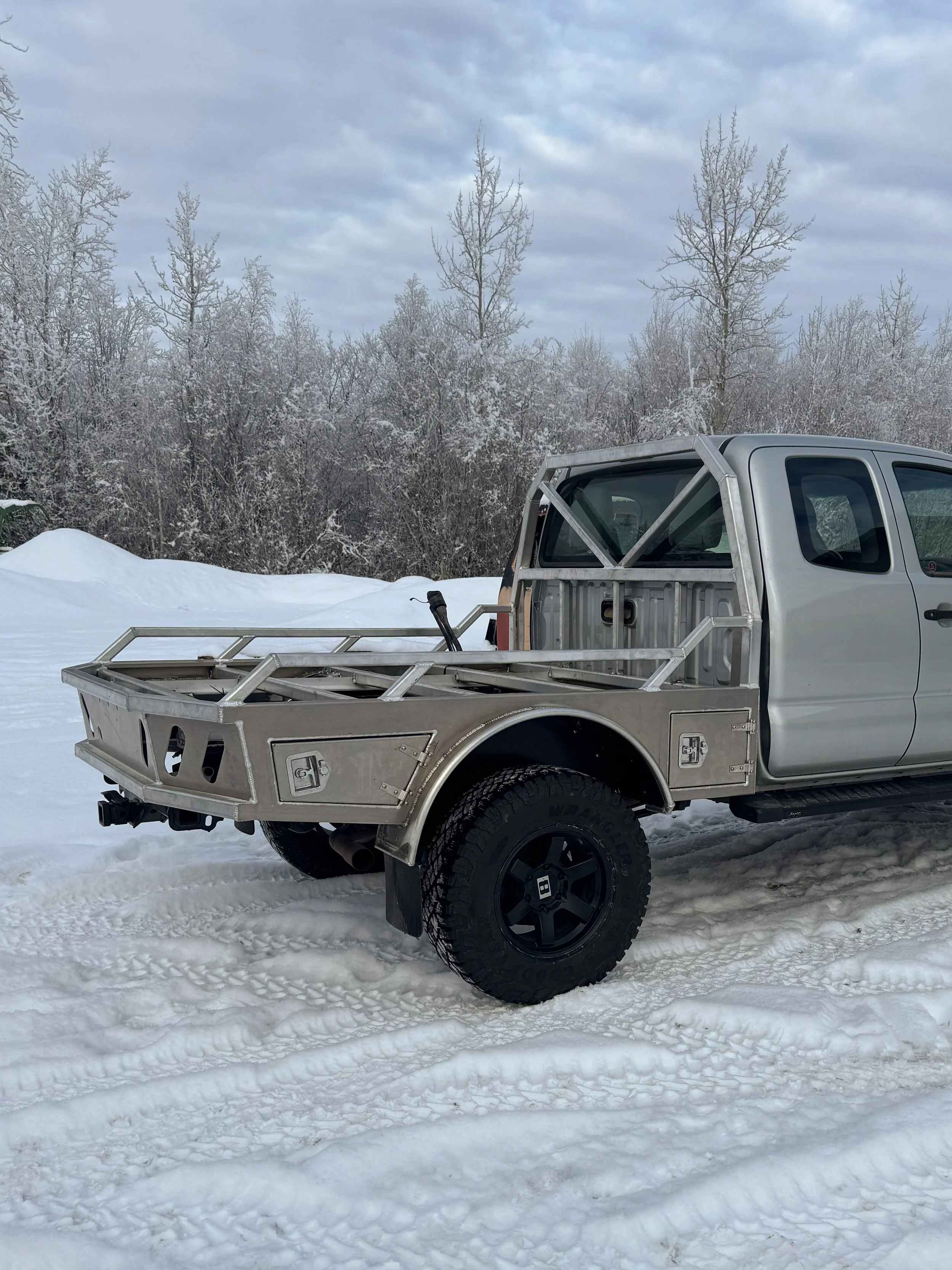 Silver pickup truck with custom built metal flat deck in snowy outdoor setting with snow-covered trees and cloudy sky in the background.