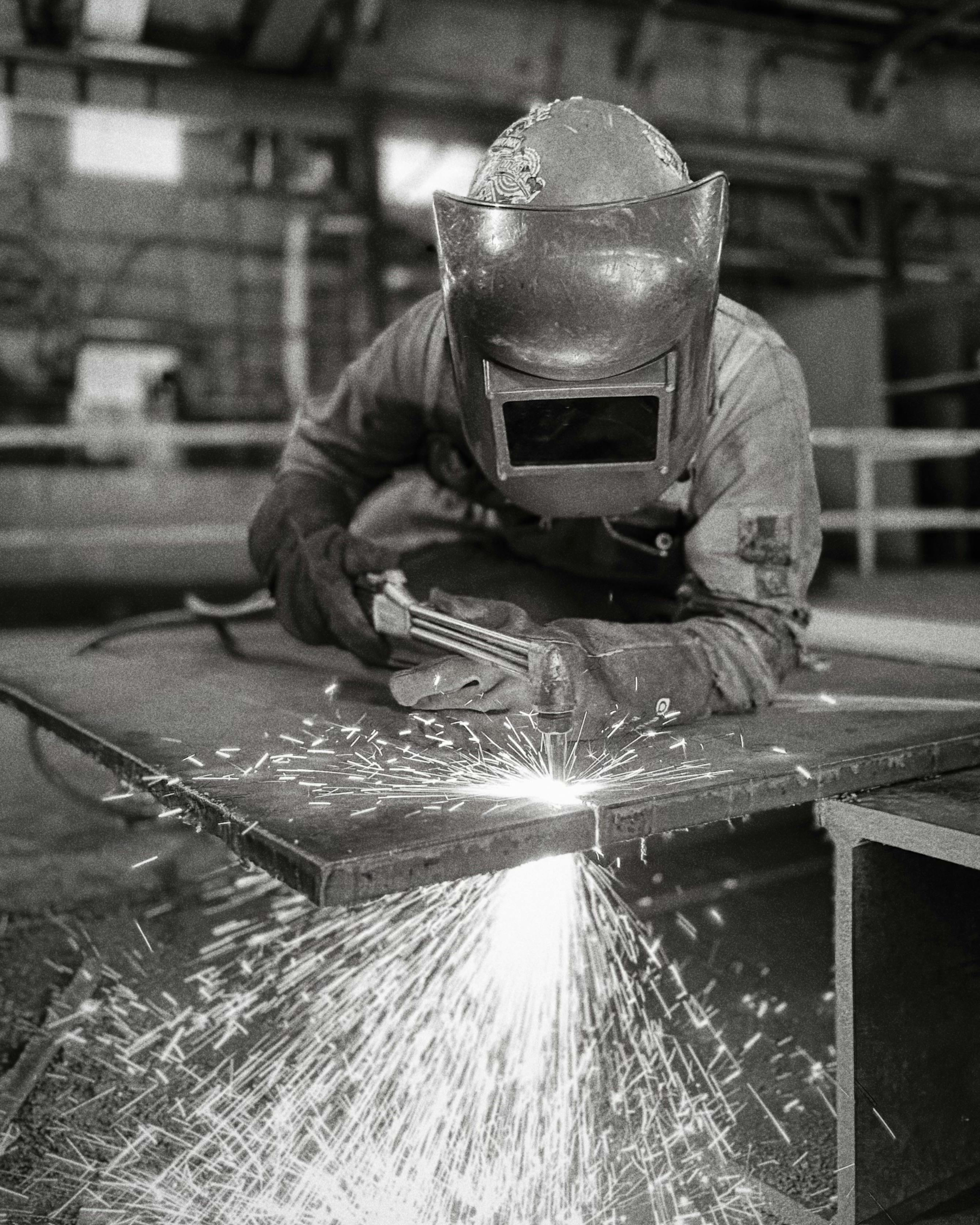 A person welding metal while wearing a welding helmet, gloves, and work clothes in a workshop.