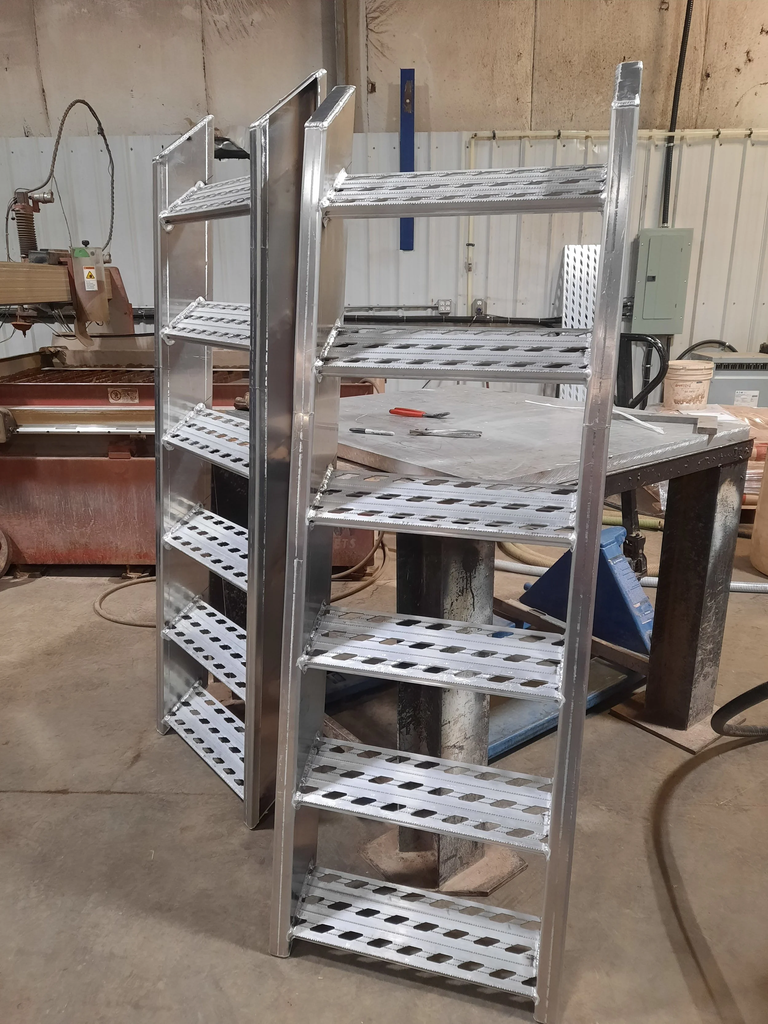 A metal steps or stairs unit with multiple grippy steps in a workshop, with tools and equipment on a workbench in the background.