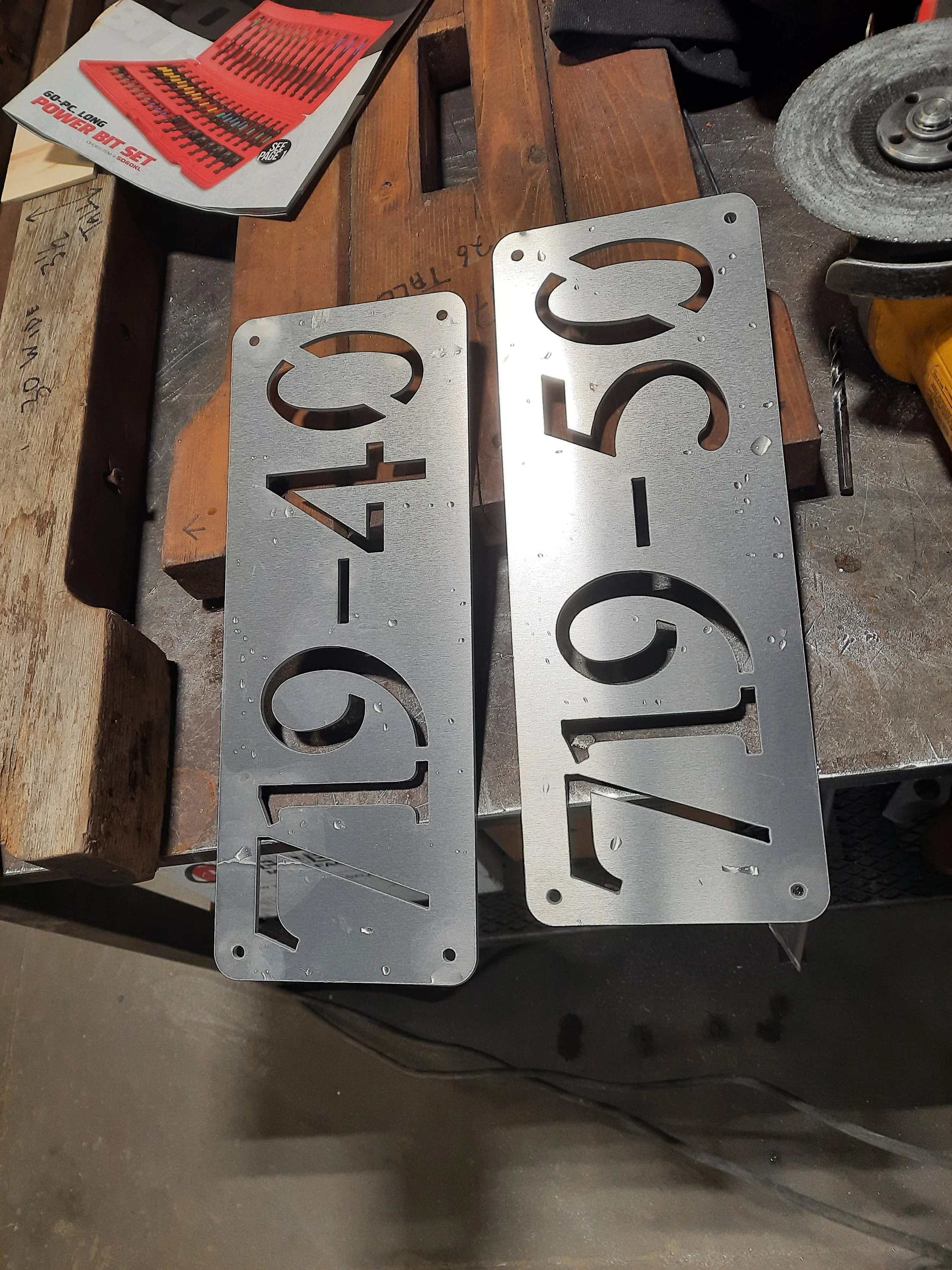 Two metal address sign plates with numbers on a workbench surrounded by tools and materials.