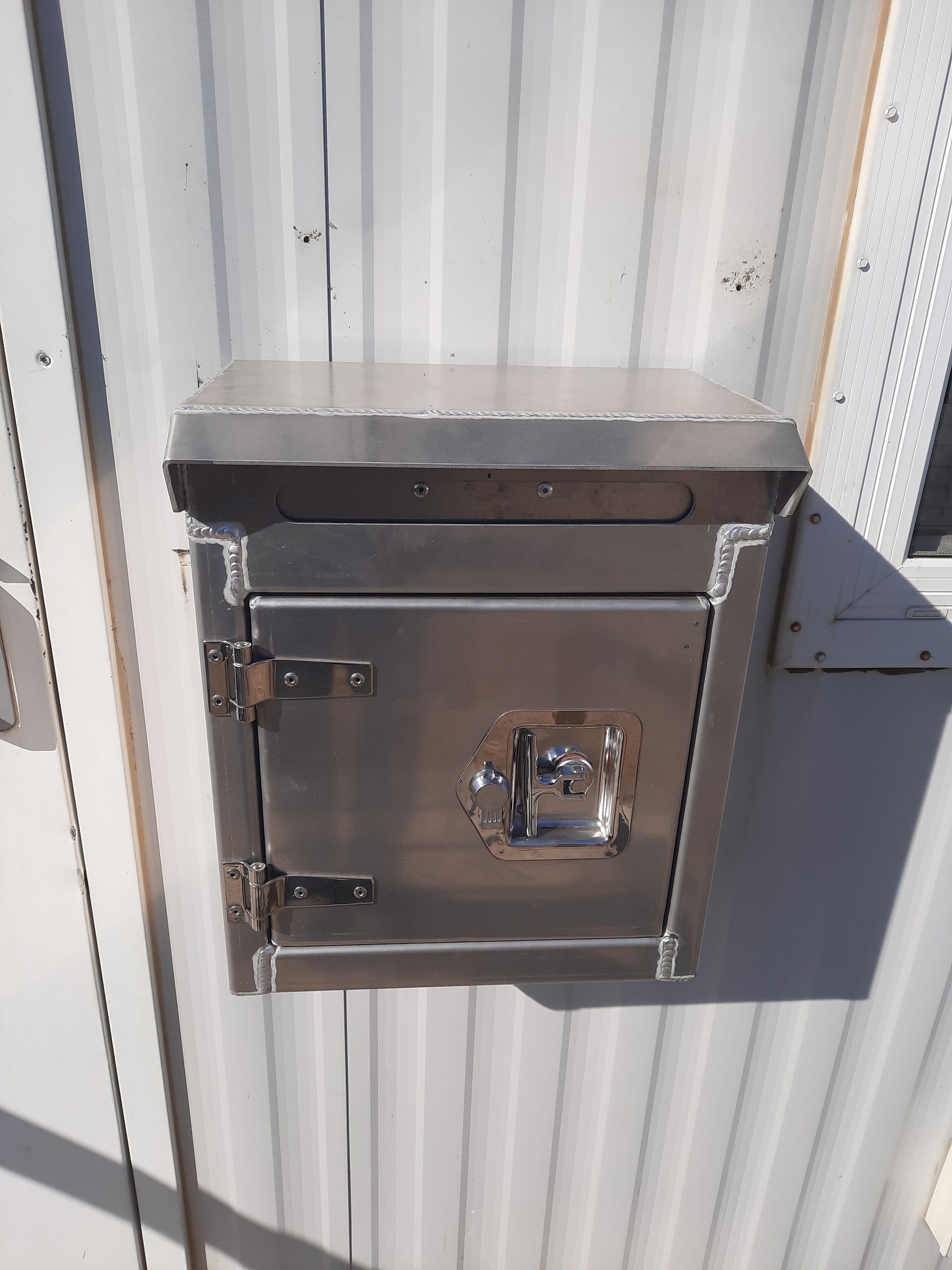 A stainless steel mailbox mounted on a white corrugated metal wall.