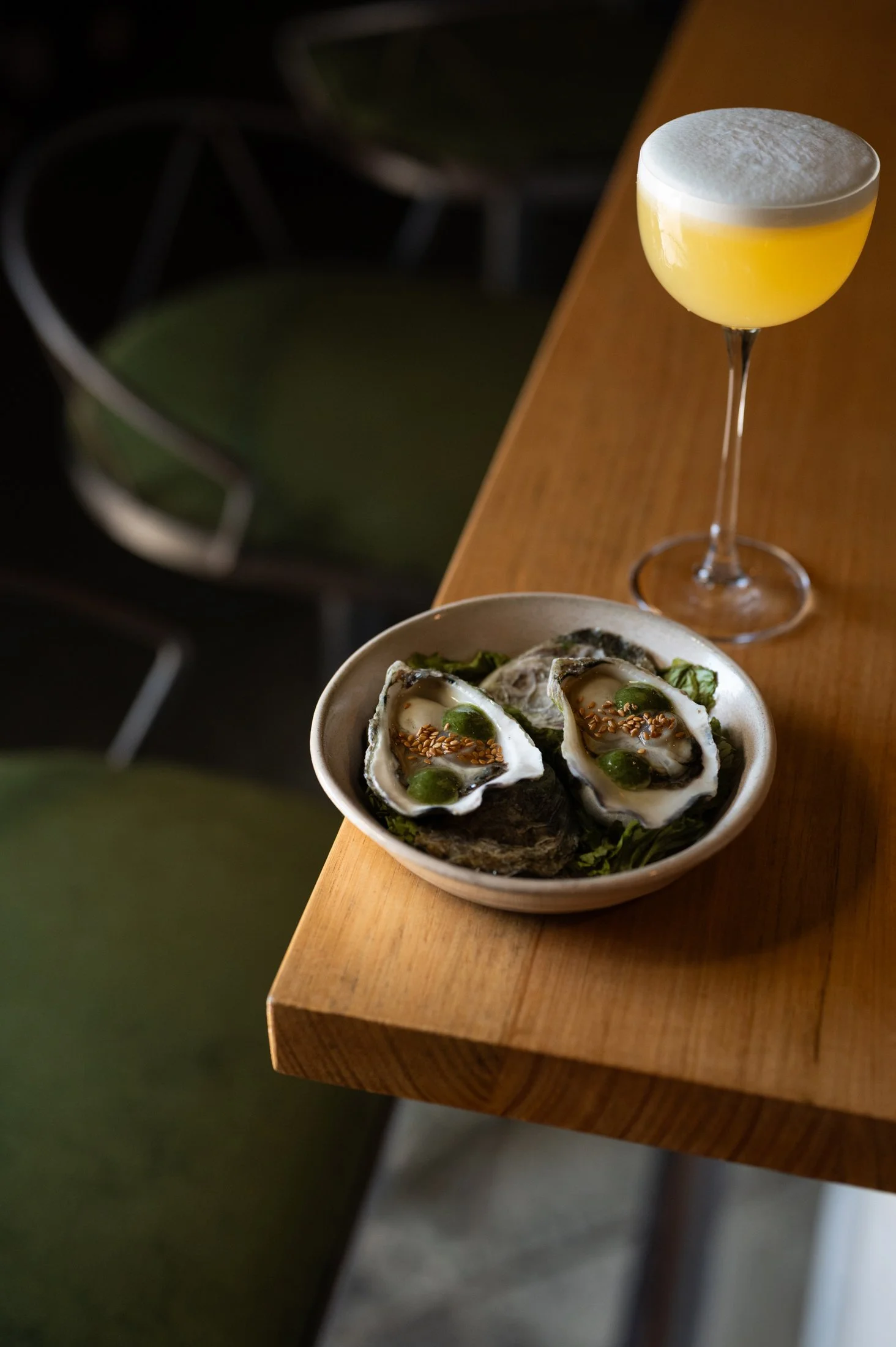 A wooden table with a bowl of oysters topped with green sauce and sesame seeds, and a cocktail with a frothy white top.
