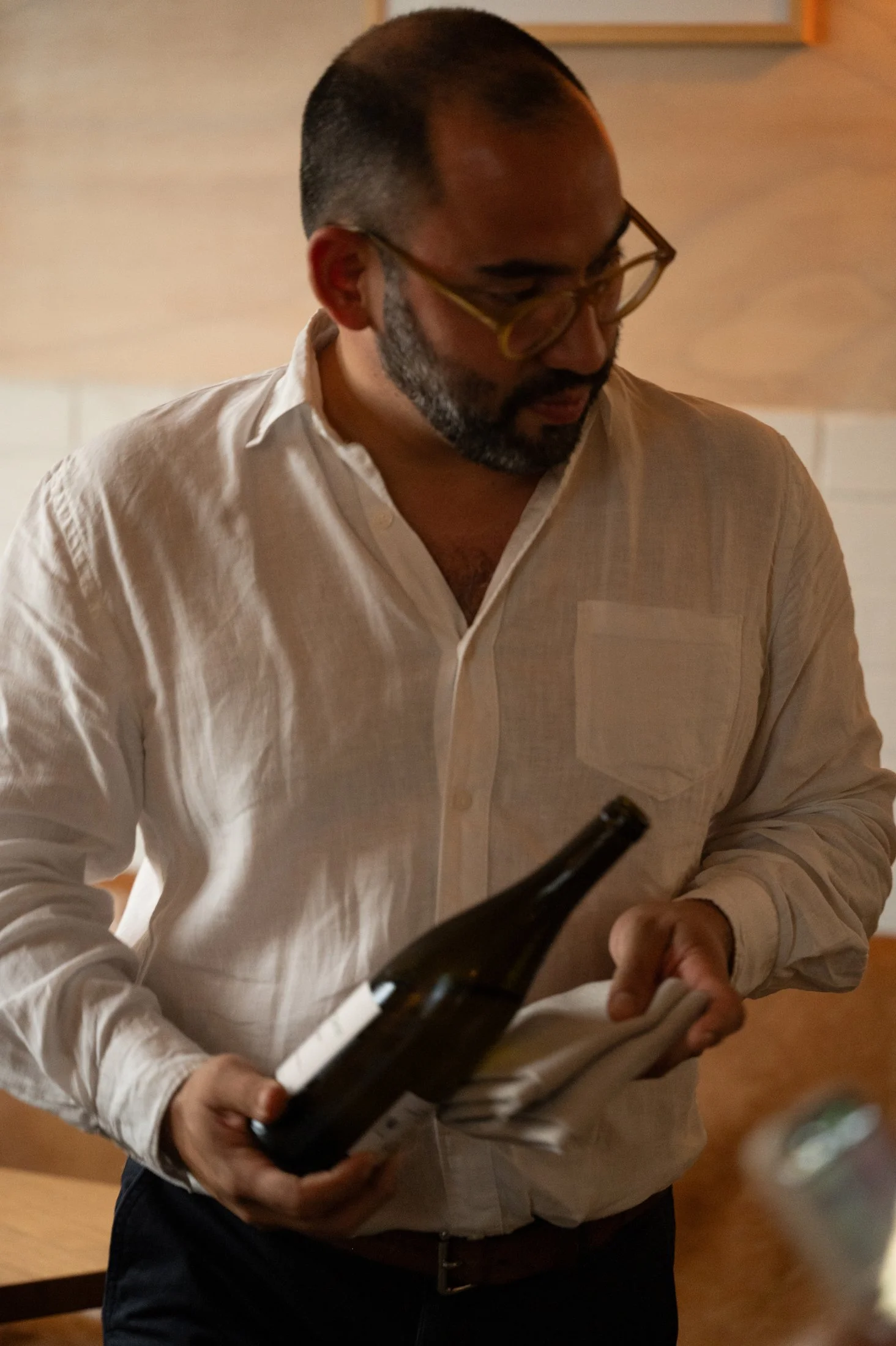 A man wearing glasses and a beige button-up shirt holding a bottle of wine and reading a book in an indoor setting.