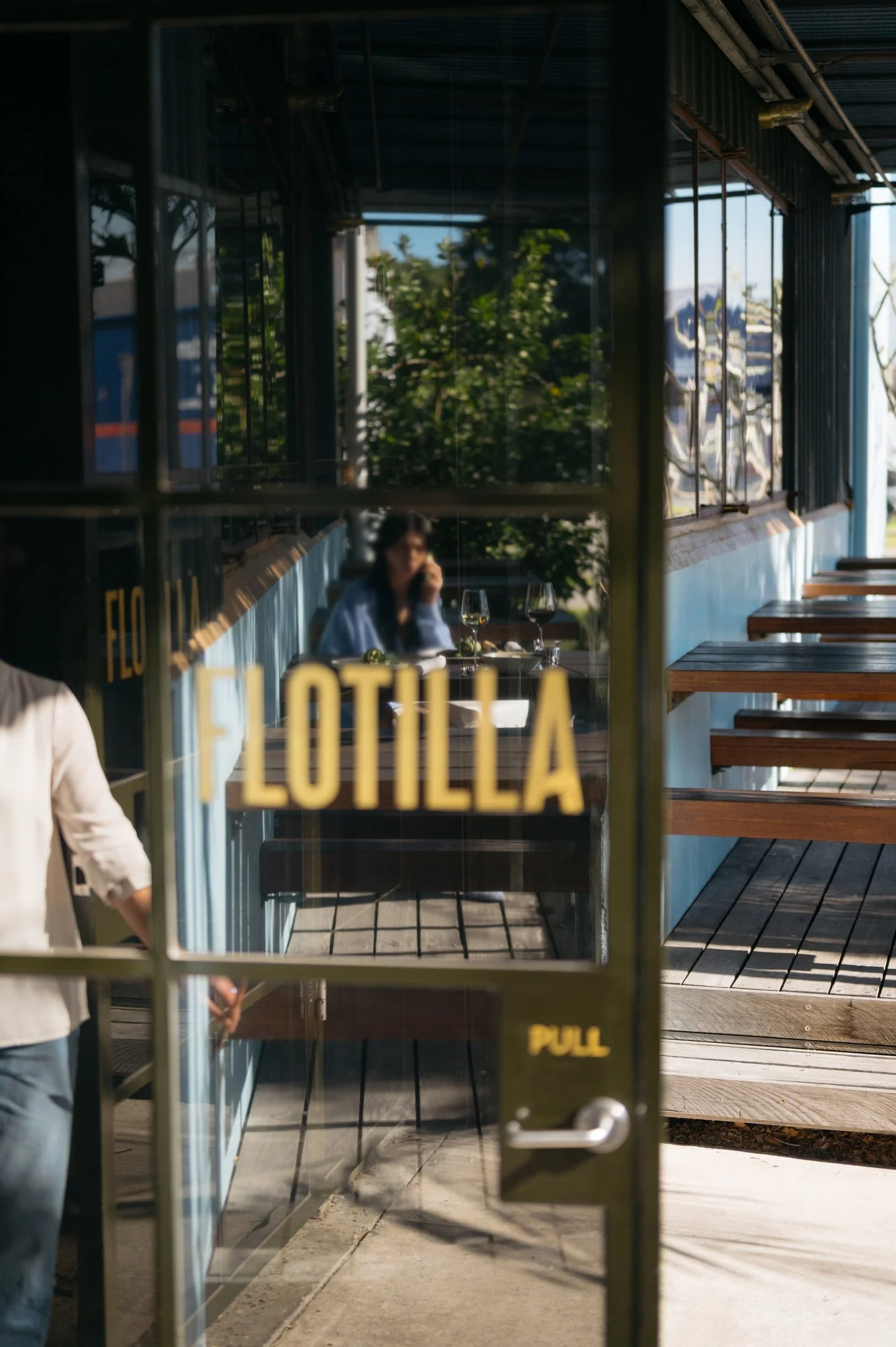 A person sitting outside at a restaurant table with wine glasses, viewed through a glass door that says 'Pull' in yellow letters, with sunlight and trees in the background.