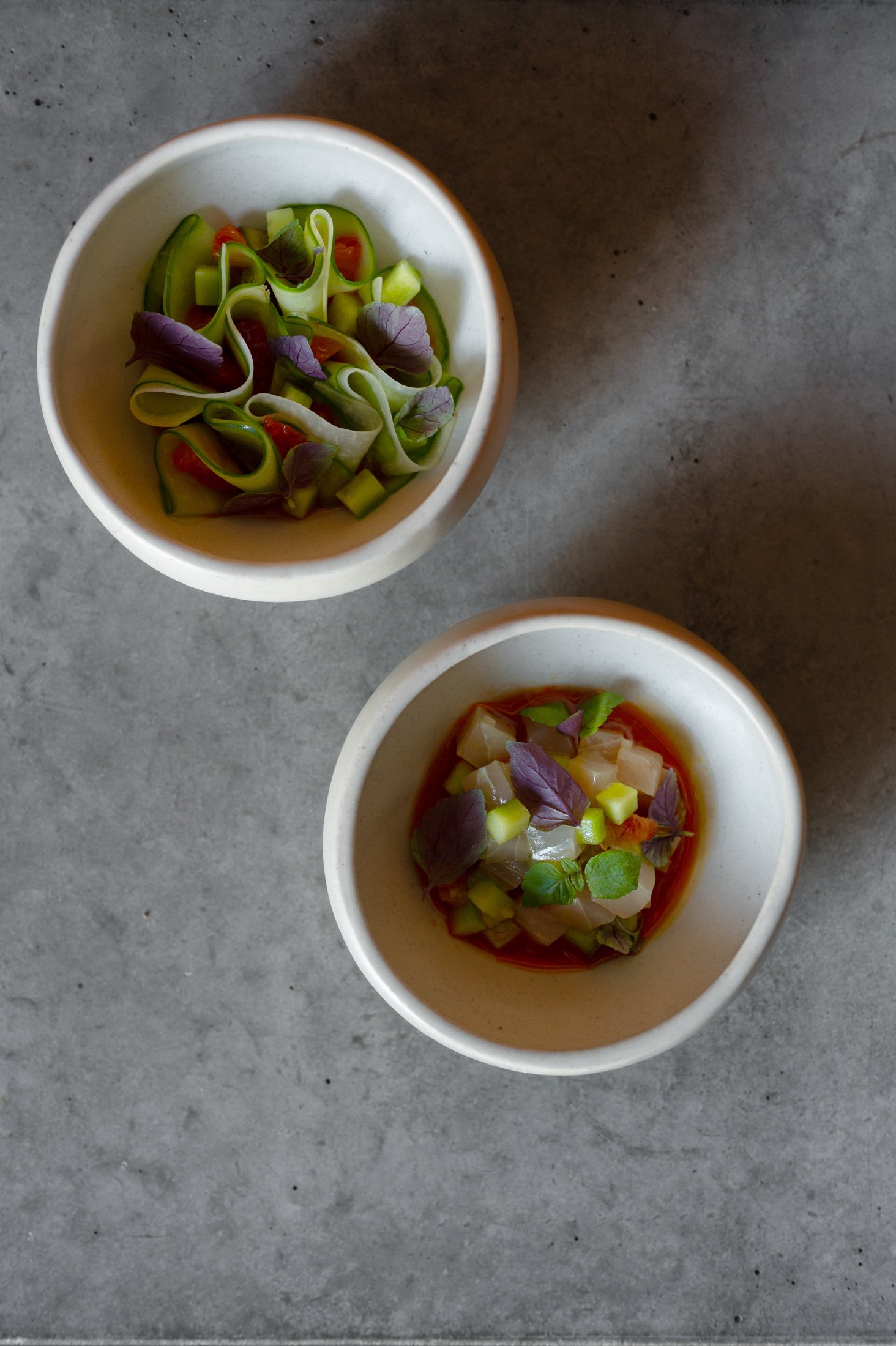Two small bowls of soup with diced vegetables and herbs on a gray surface.