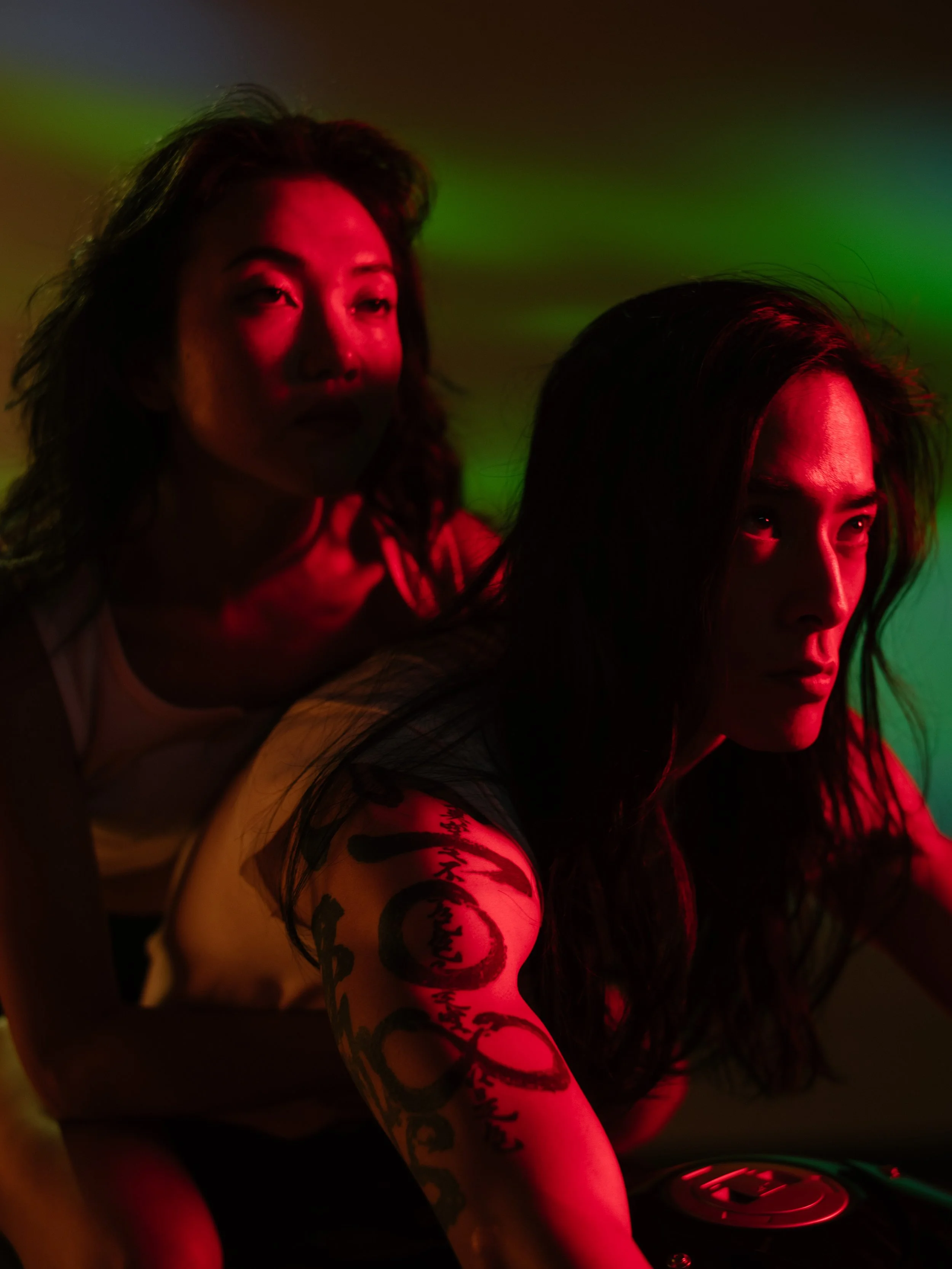 Two women with dark hair in a dimly lit room with colorful lighting. One woman is in the foreground with a prominent tattoo on her arm, and the other woman is in the background with her face partially illuminated.