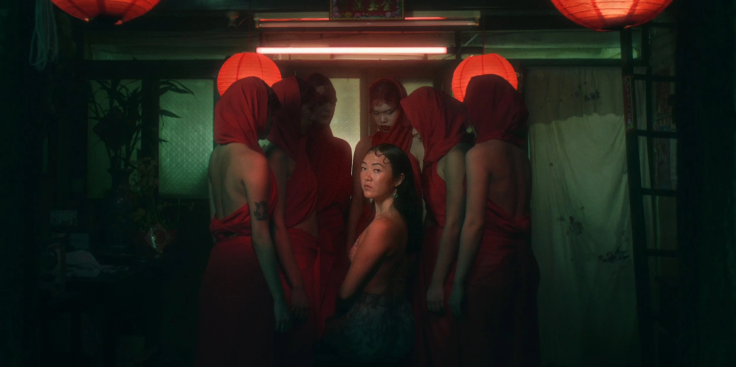 A woman sitting on the floor surrounded by six other women dressed in red, with red lanterns and green lighting in the background.