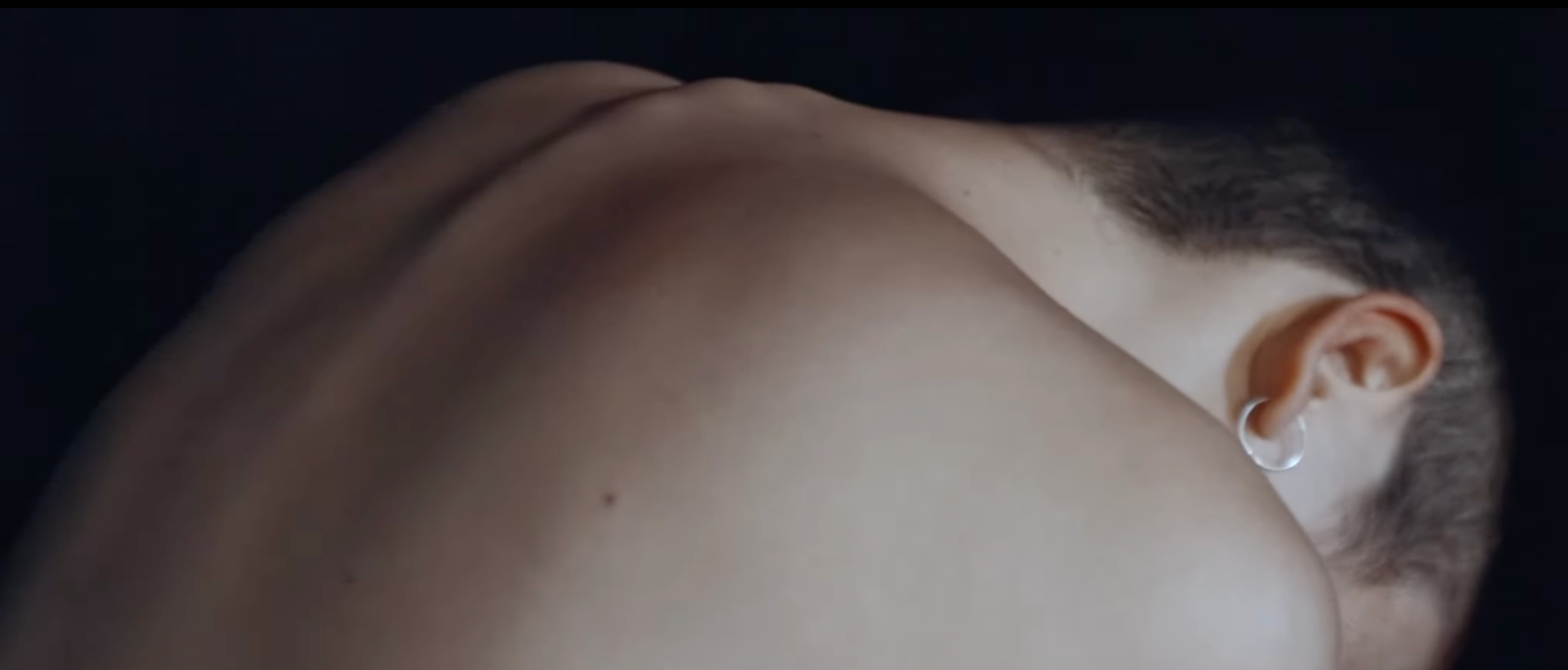 Close-up of a person's shoulder and neck with short hair, wearing a hoop earring, against a dark background.