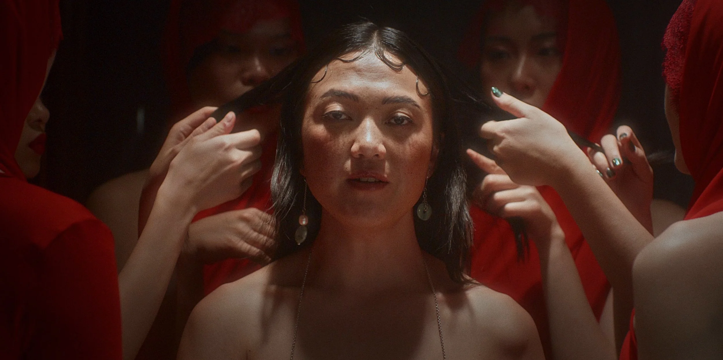 A woman with dark hair and earrings is surrounded by several women in red clothing who are touching her hair.