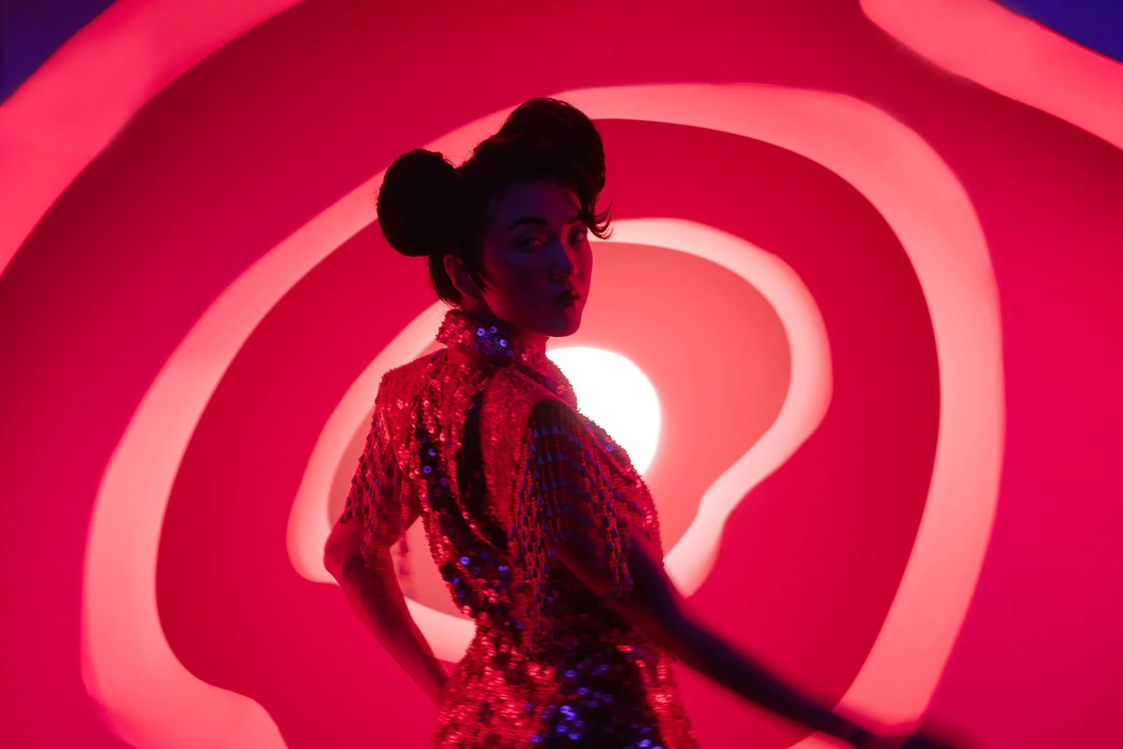 A woman in a shiny, sequined outfit stands in front of a vibrant, pink and red neon abstract background with swirling patterns, creating a dramatic and artistic scene.