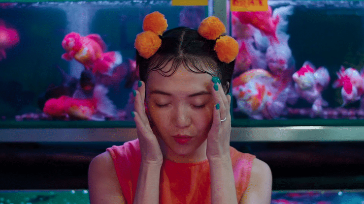 A young woman with dark hair in buns decorated with orange pom-poms, closing her eyes with her hands on her face in front of an aquarium filled with colorful fishes.