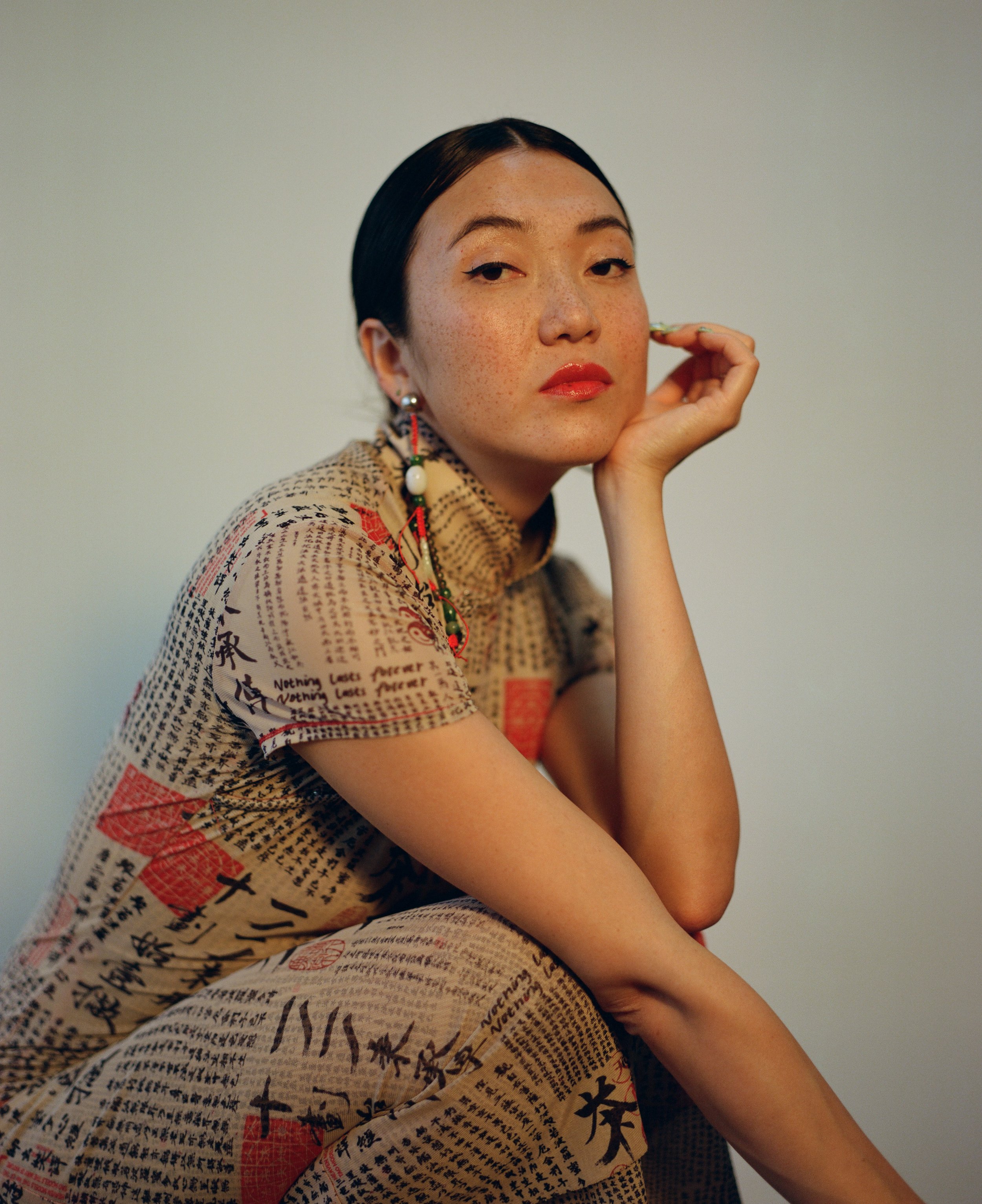 A woman with short dark hair, light makeup, red lipstick, wearing a patterned outfit with Japanese characters, sitting with her chin resting on her hand, looking at the camera.