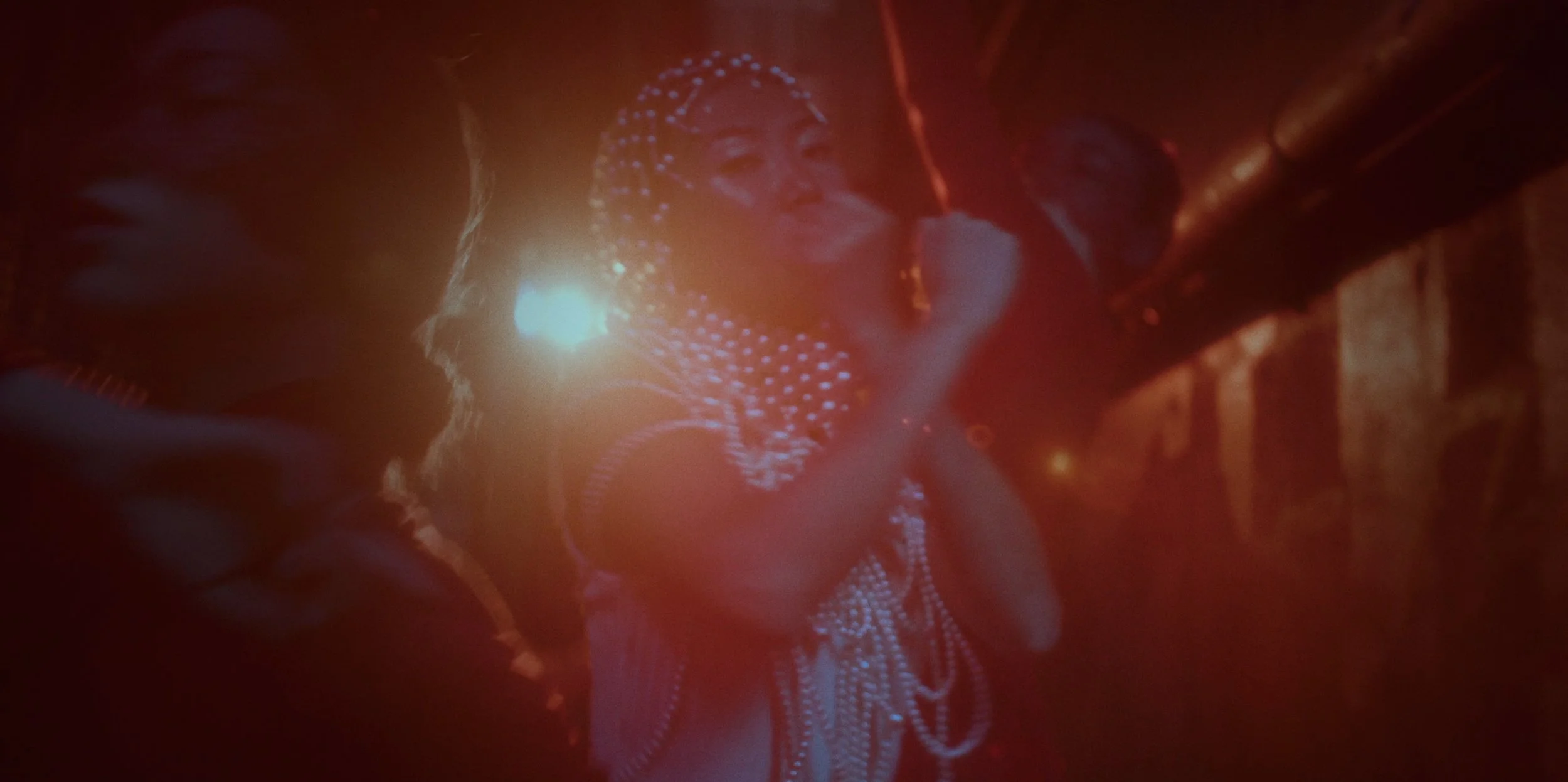 A woman wearing rhinestone-studded clothing and head covering, speaking into a microphone on stage, with red and orange lighting creating a vibrant atmosphere.