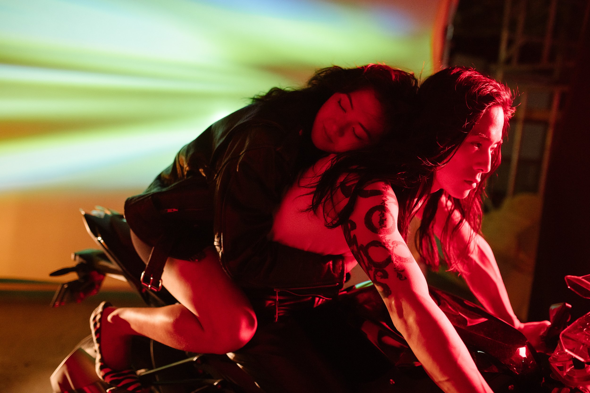 Two women riding a motorcycle in a dimly lit environment with colorful lights, one in front and the other riding behind, leaning forward with an intense focus.