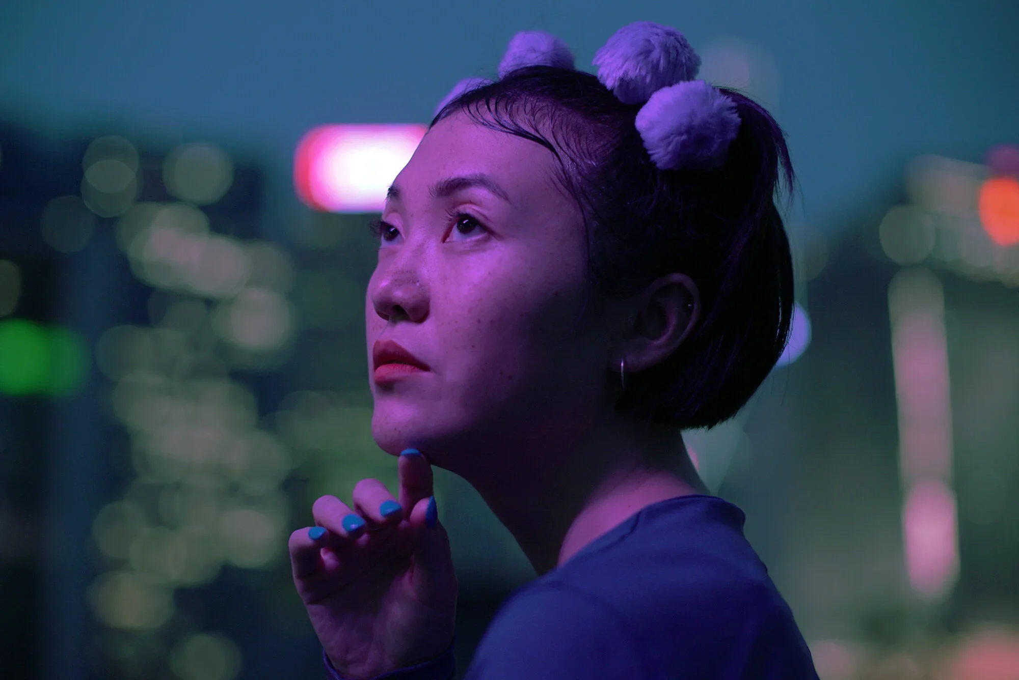 A young woman with short dark hair styled with fluffy hair accessories, illuminated by purple and blue city lights at night, with a contemplative expression and her hand gently touching her chin.