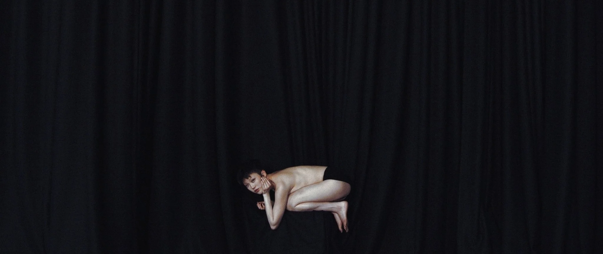 A woman with short haircut lying on her side in a fetal position on a black stage or backdrop.