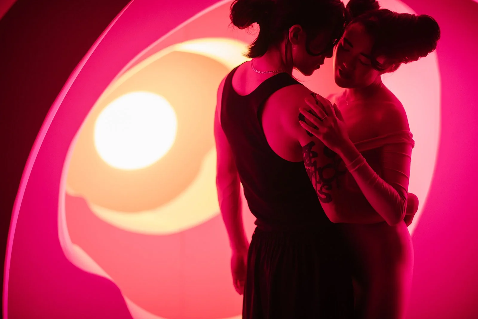 Two people embracing in front of a neon pink and yellow light with a circle shape, one wearing a sleeveless top and the other wearing a long-sleeve garment, both with dark hair, sharing a tender moment.