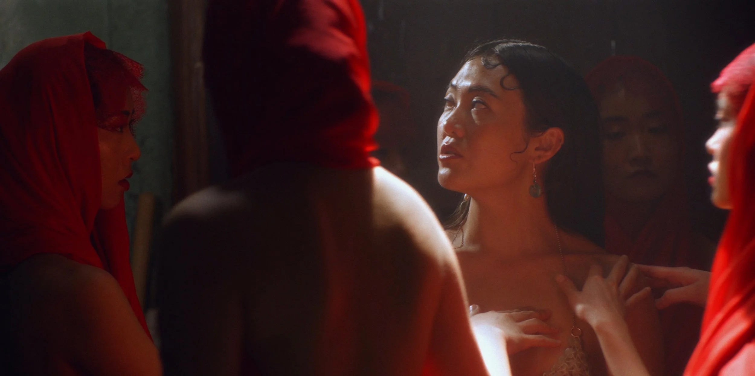 A woman with dark hair and jewelry, looking upward with a serious expression, surrounded by women in red hoods in a dark setting.