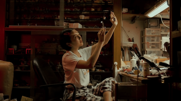 A young girl with curlers in her hair sitting in a cluttered workshop or craft room, holding up a small object and inspecting it closely while sitting on a chair.