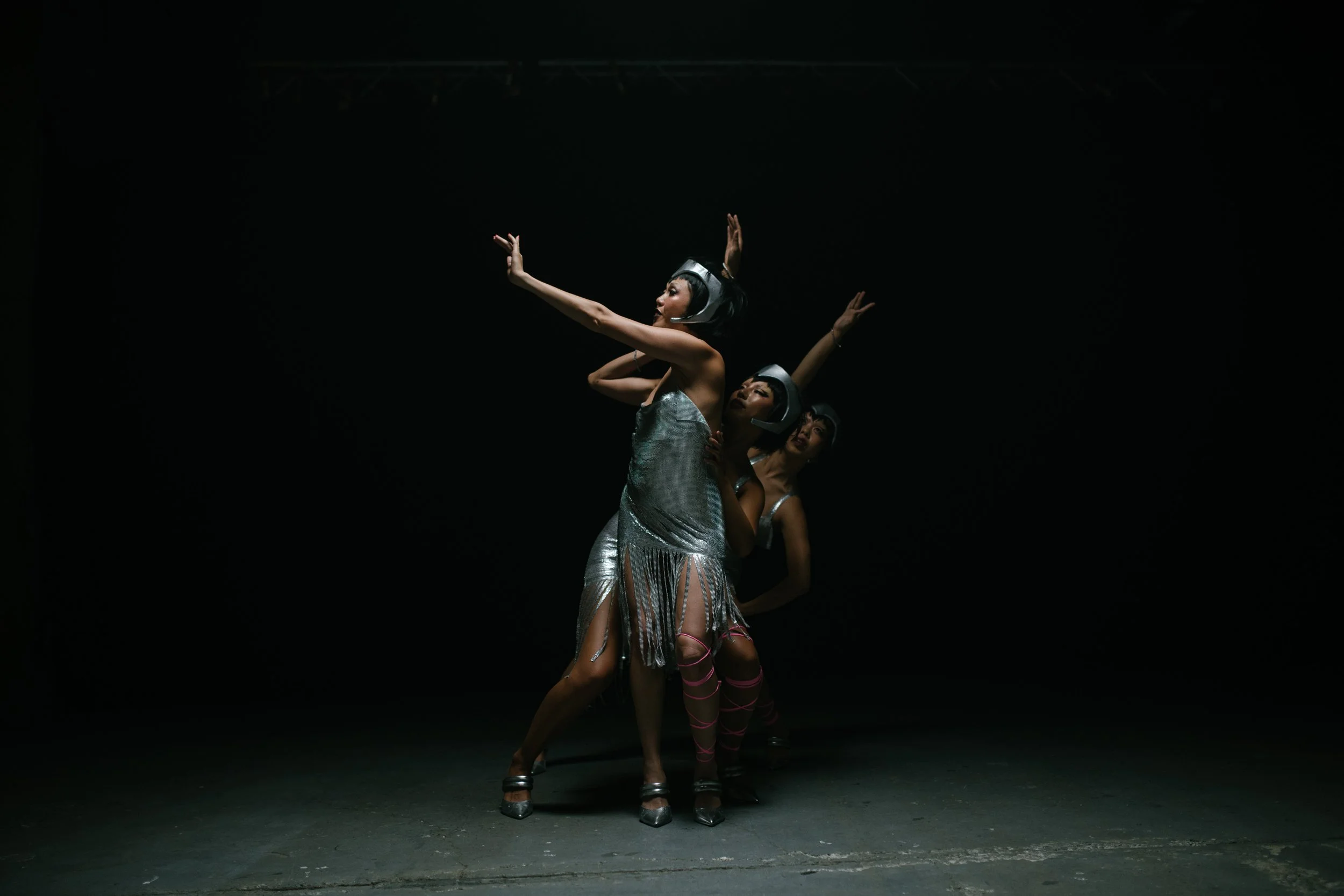 Three women dance in metallic costumes with futuristic helmets on a dark stage, performing a choreographed routine.