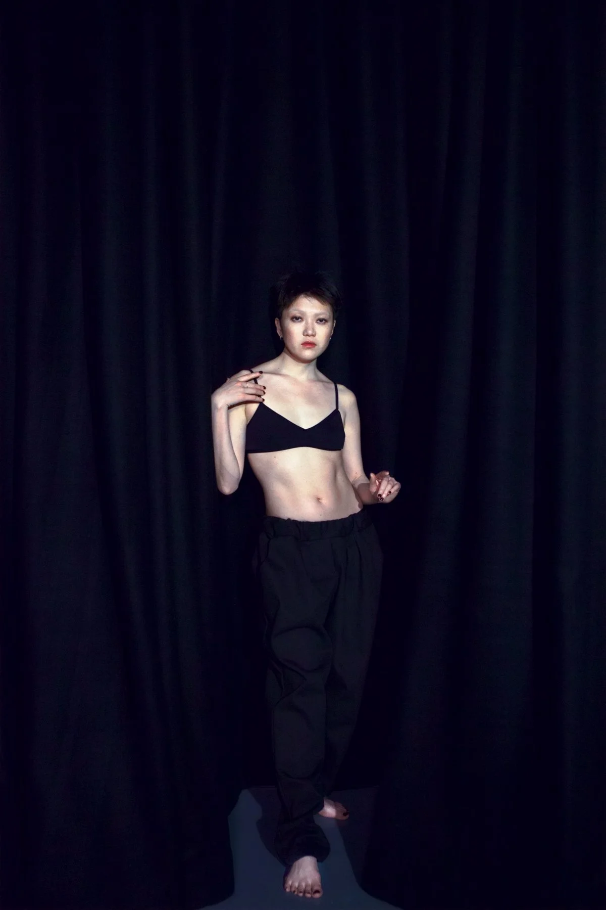 A young woman with short dark hair stands in front of a black curtain, wearing a black crop top and black pants, looking directly at the camera.