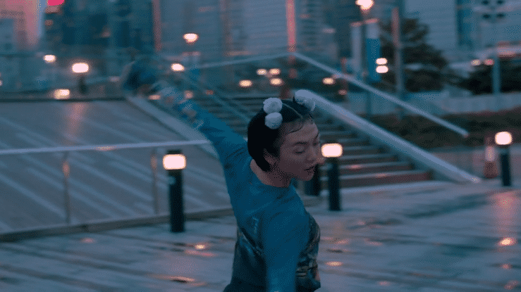 A woman dancing outdoors at dusk, with city buildings and streetlights in the background.