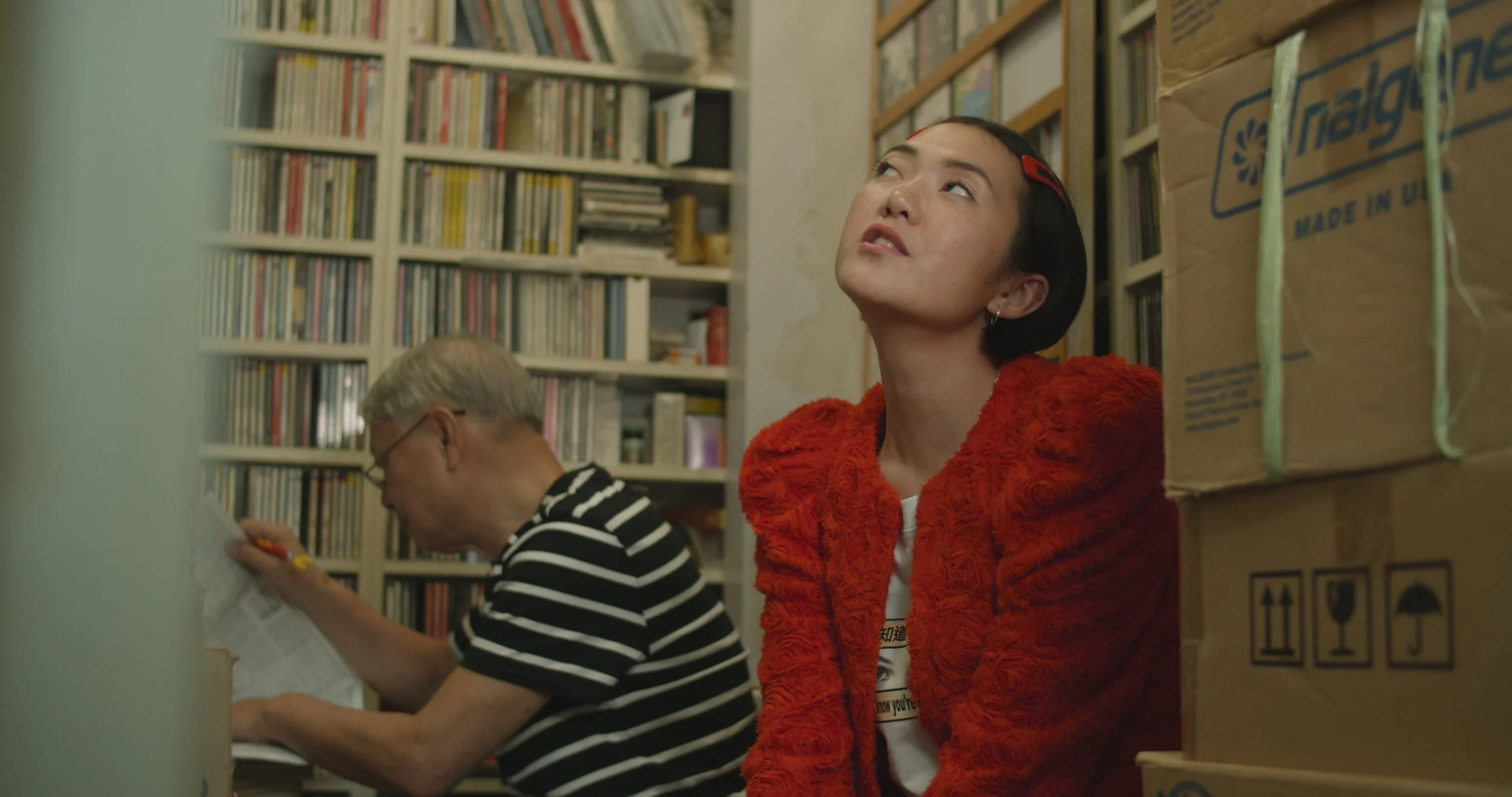 A woman with short black hair and earrings in an orange furry jacket sitting among boxes and bookshelves, looking upward with a thoughtful expression. An older man with glasses in a black and white striped shirt is reading a paper in the background.