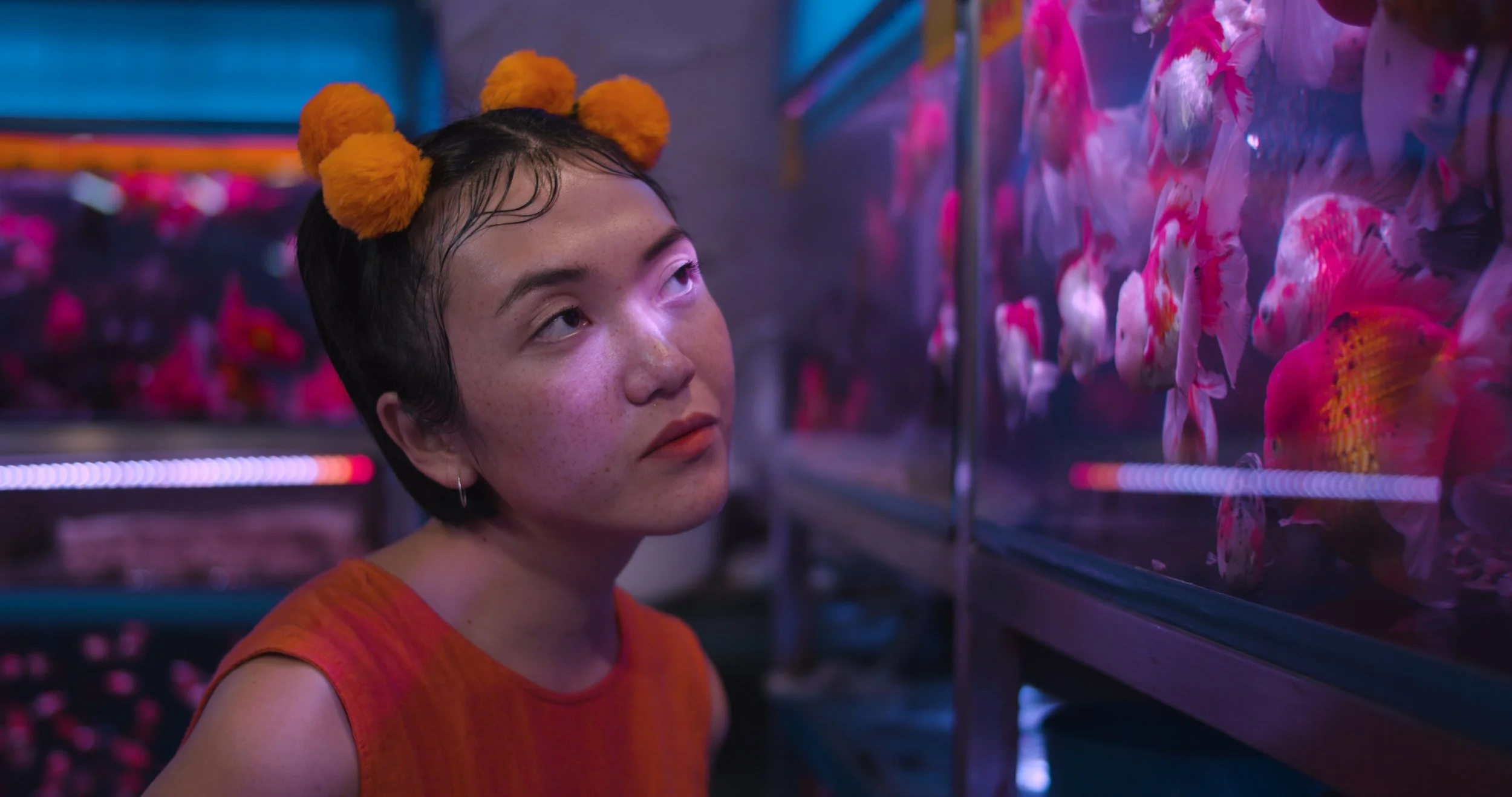 A young woman with short dark hair, freckles, and orange hair accessories is looking at a display of colorful artificial flowers in a store.