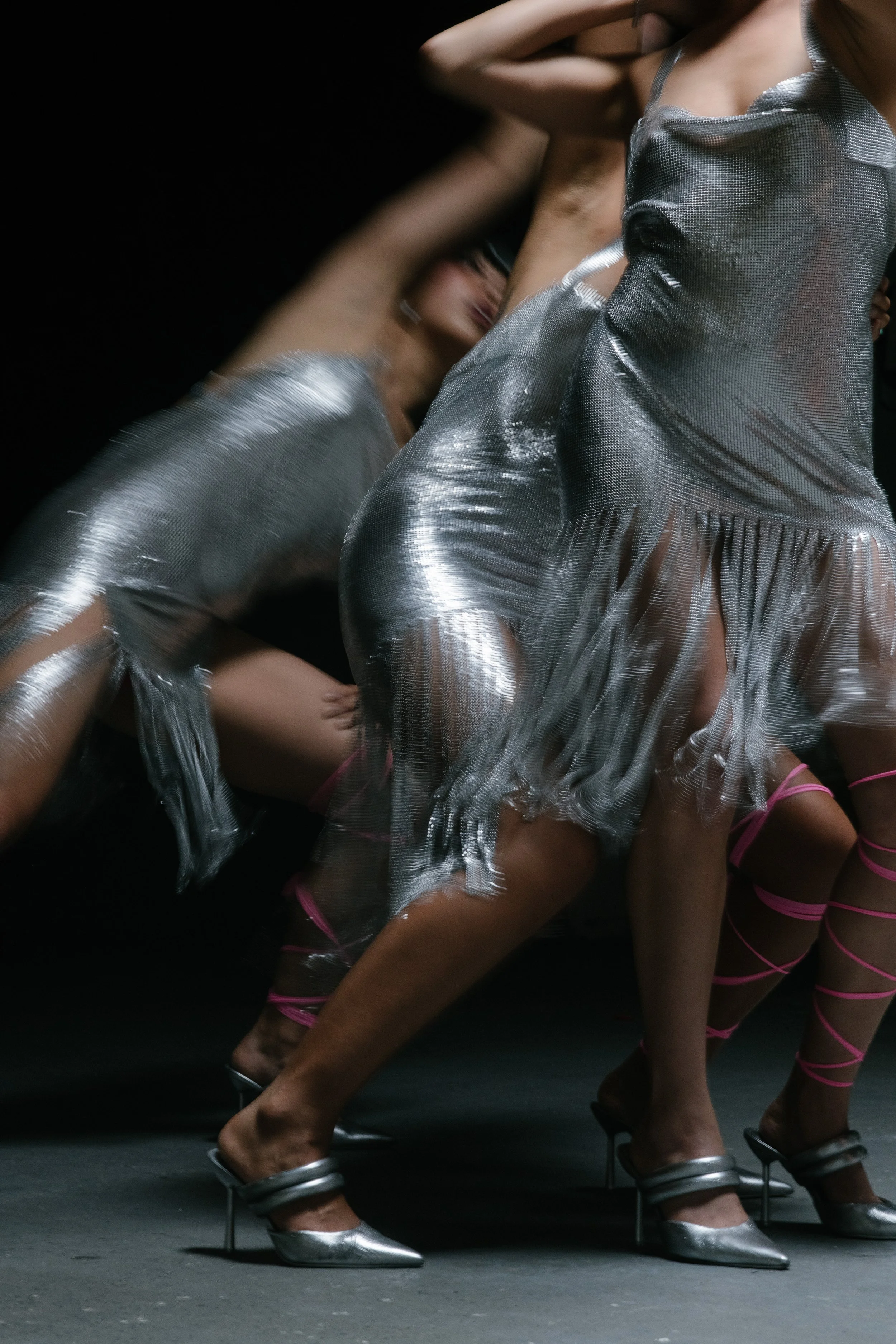 Group of women in metallic dresses and heels, crouching on the floor, some with pink lace-up shoes, in a fashion runway setting.