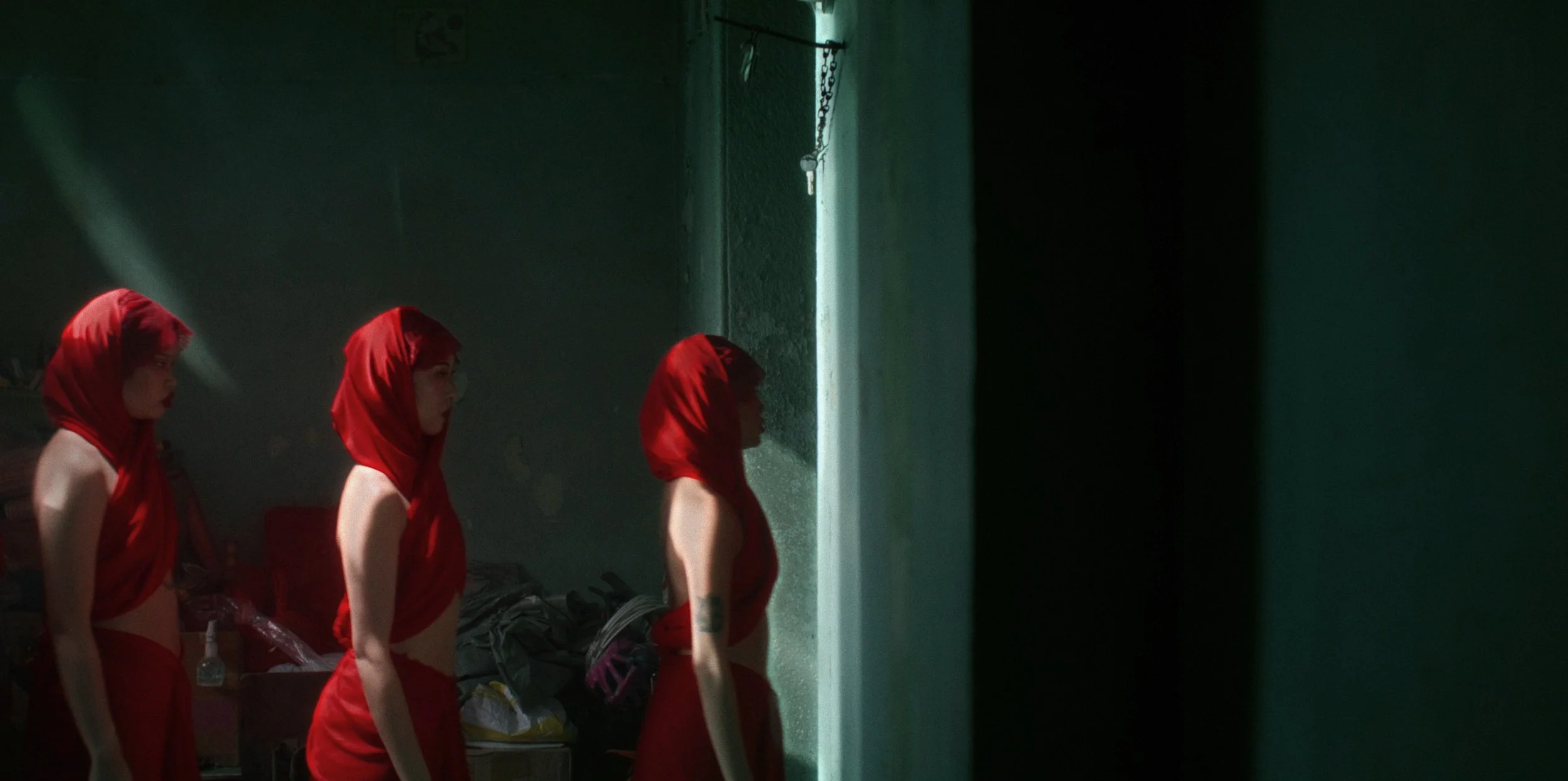 Four women with red hooded scarves, topless from the waist up, standing in profile in a dimly lit room, with a single light source casting shadows on their faces and a wall with a door and laundry in the background.