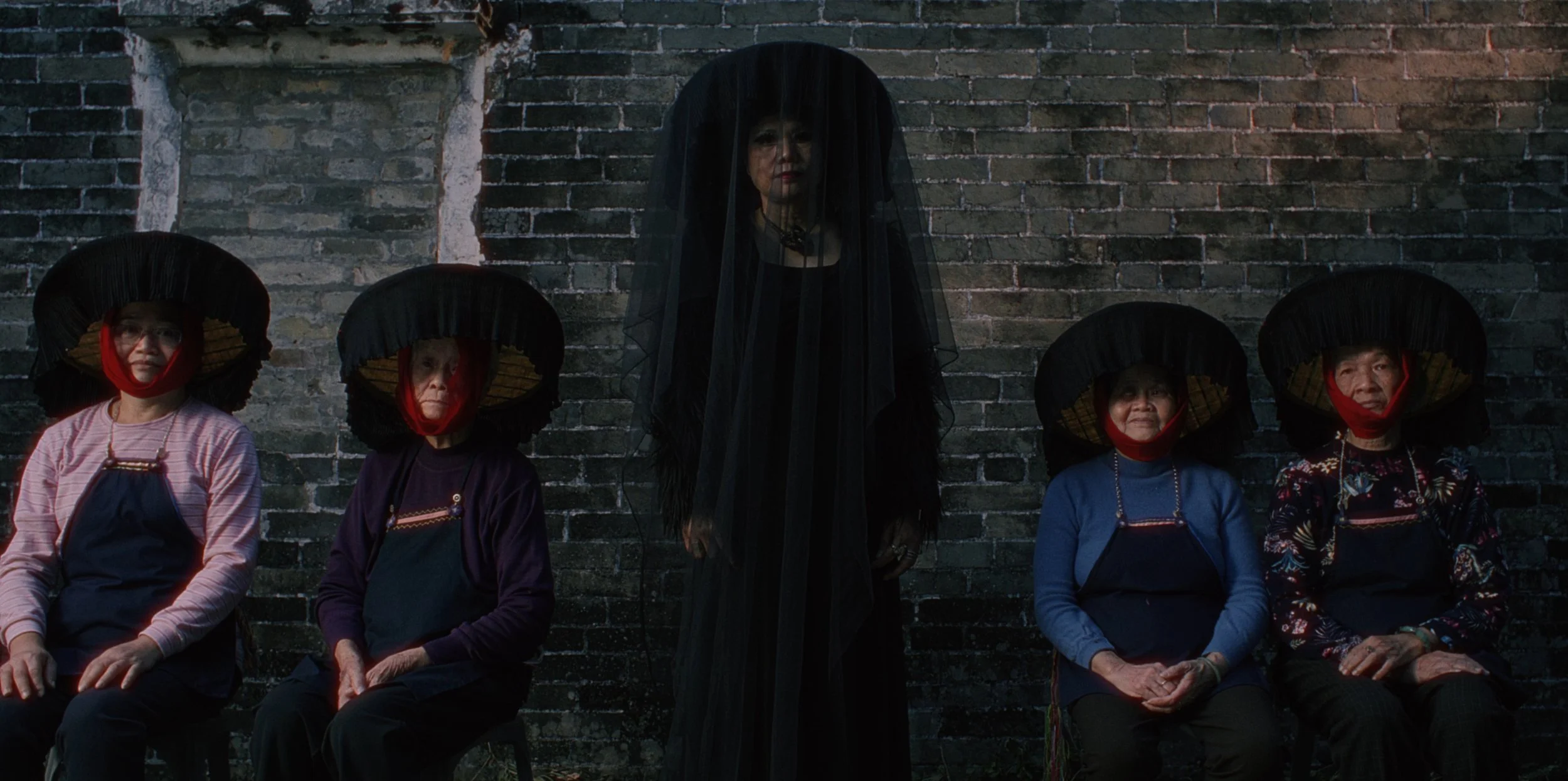 Five women sitting against a brick wall, wearing traditional Asian attire and large black hats, with one woman standing behind them dressed in all black and covered with a black veil.