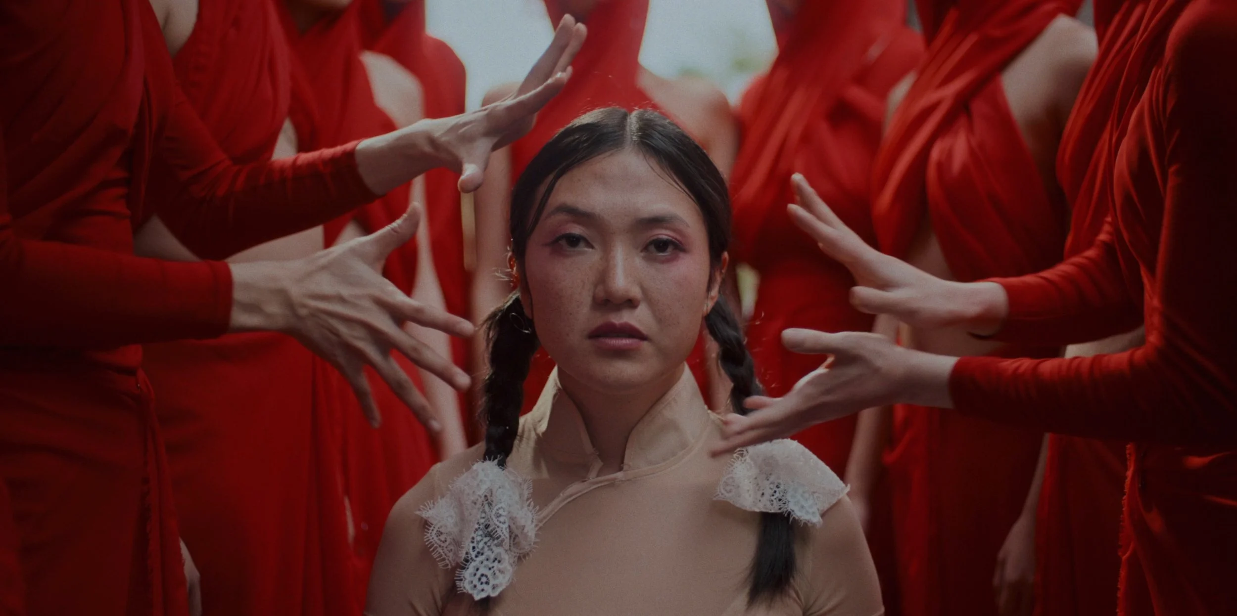 A young woman with braided hair and freckles is sitting amidst multiple people wearing red clothing. The people surrounding her are reaching out towards her with their hands, forming a frame around her face.