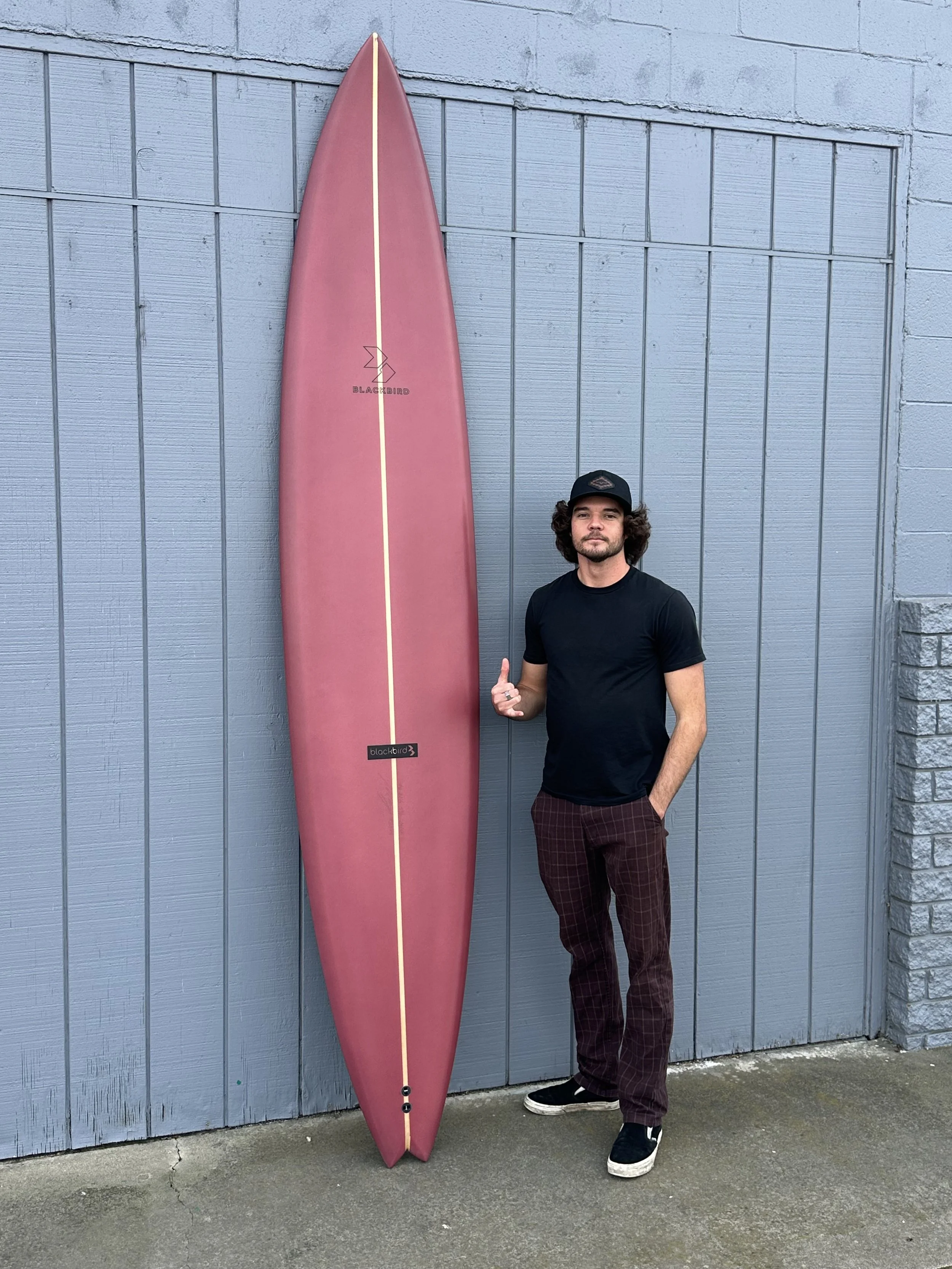 A man with curly hair and a beard, wearing a black cap, black t-shirt, plaid pants, and sneakers, standing next to a pink surfboard with a logo on it against a gray wooden fence.