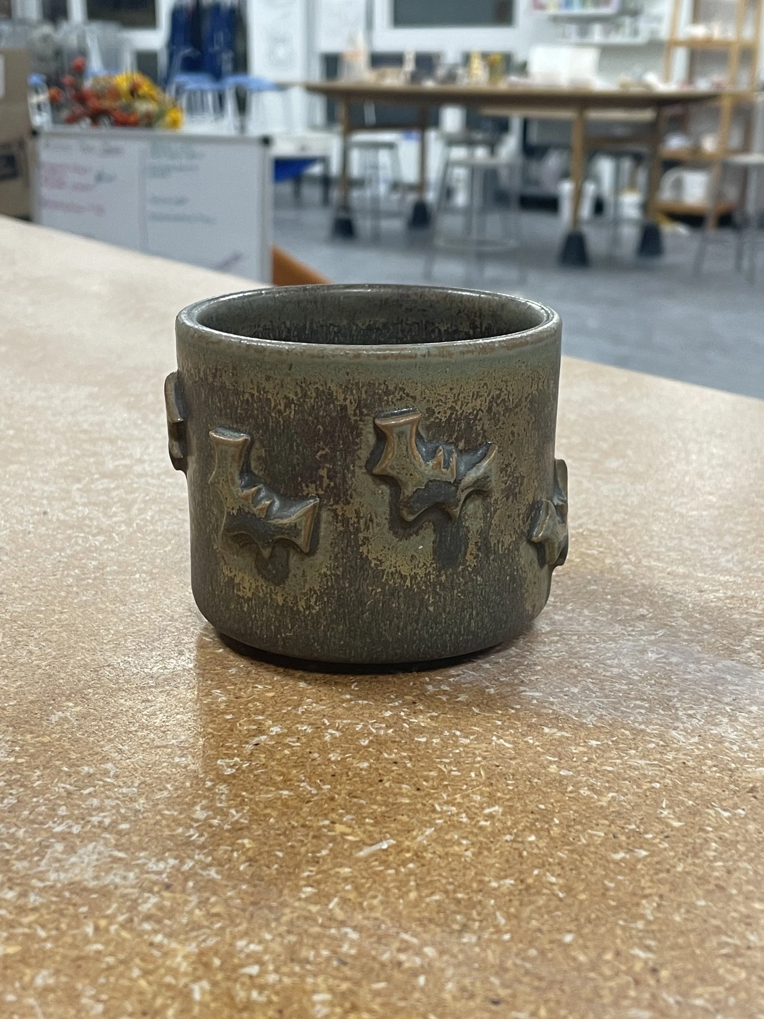A small ceramic cup with bat decorations on a light brown table, with a blurred room containing tables and chairs in the background.