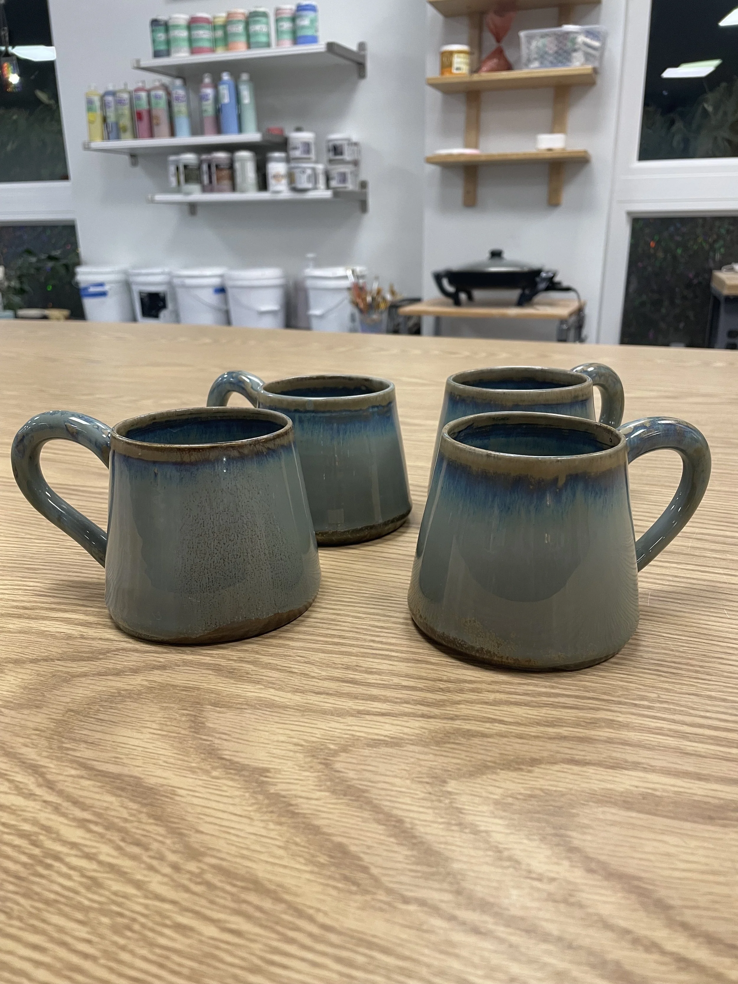 Four ceramic coffee mugs placed on a wooden table in a ceramics studio or workshop, with shelves of supplies and a window in the background.