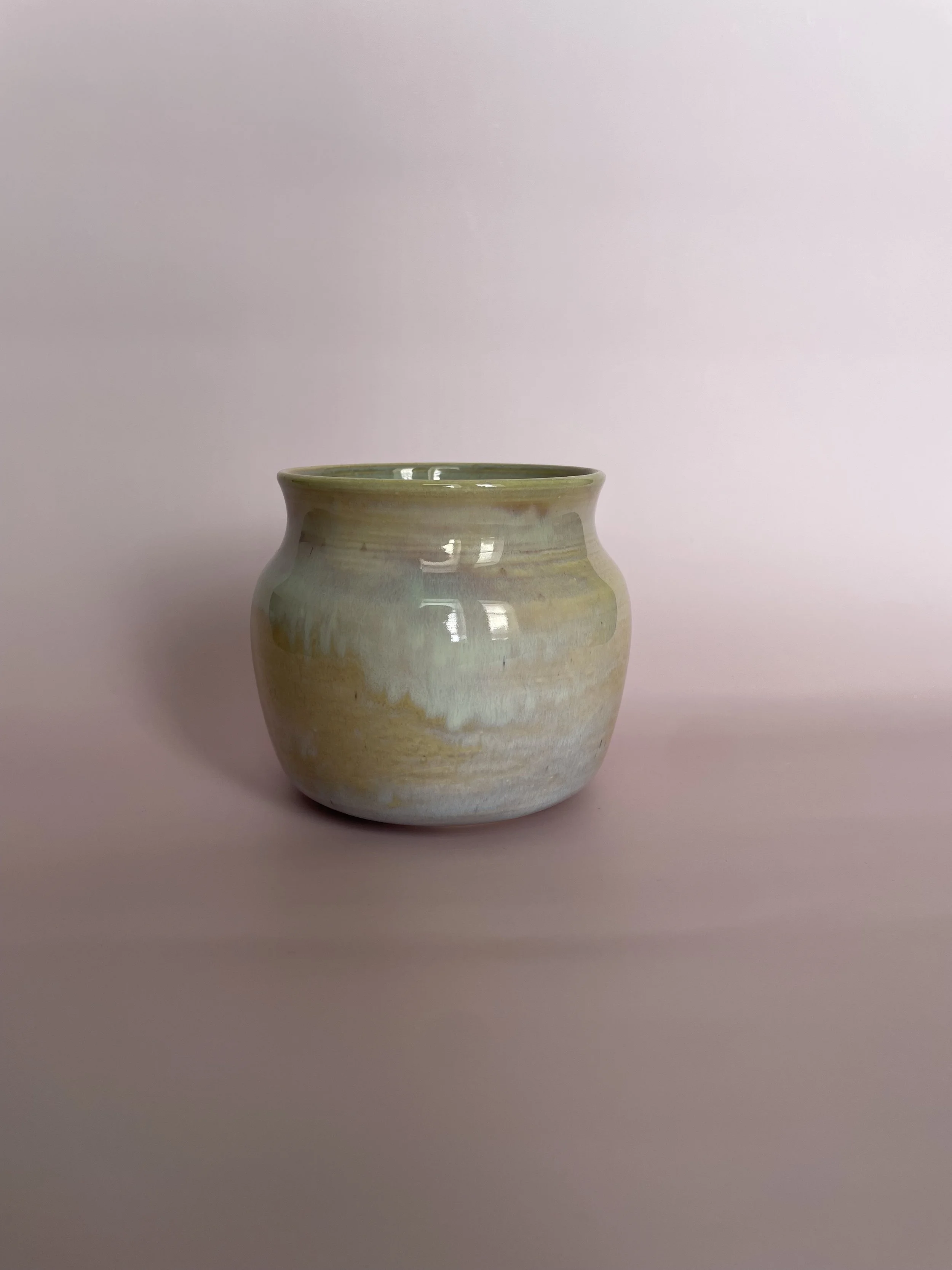 A ceramic vase with a beige and green glaze, placed on a pink surface with a matching pink background.