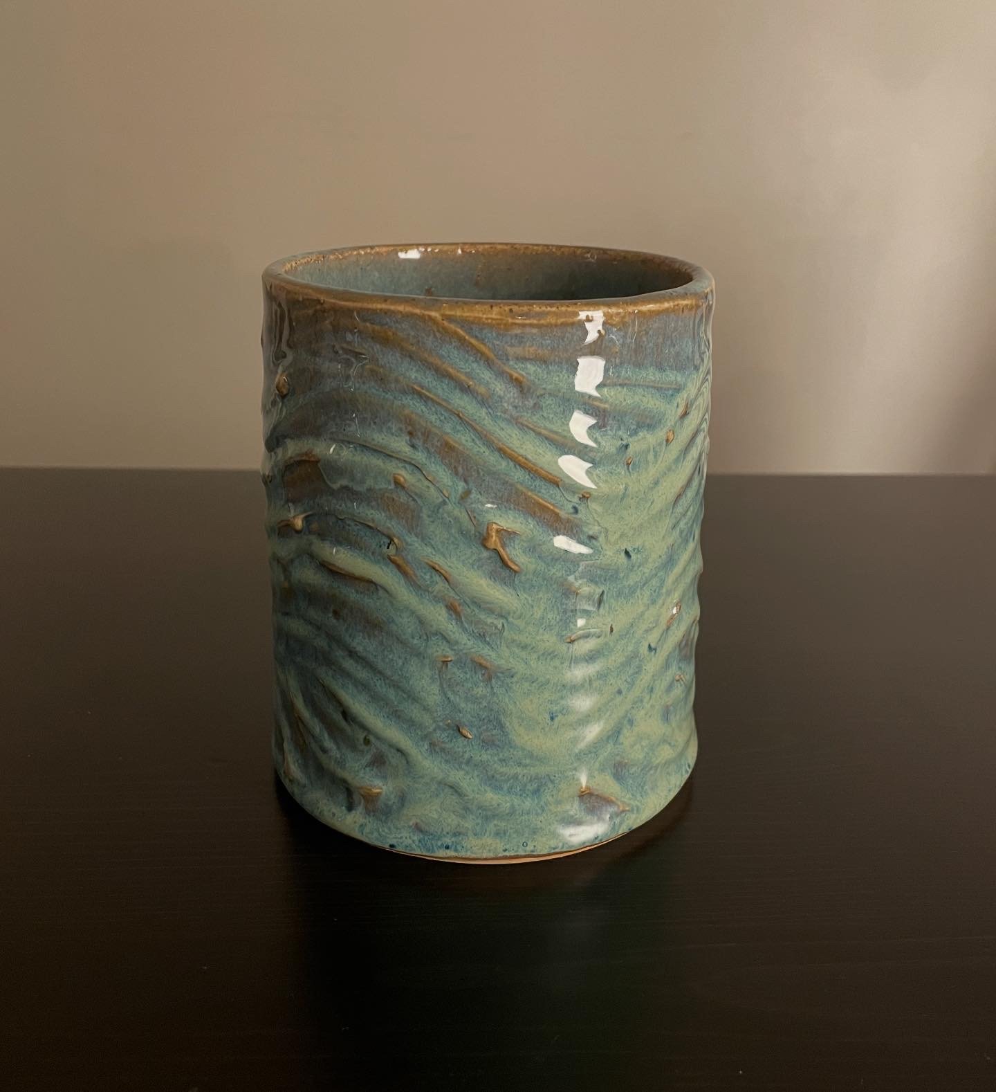 A ceramic vase with a textured, swirling pattern in shades of blue and green, sitting on a black surface against a plain wall.