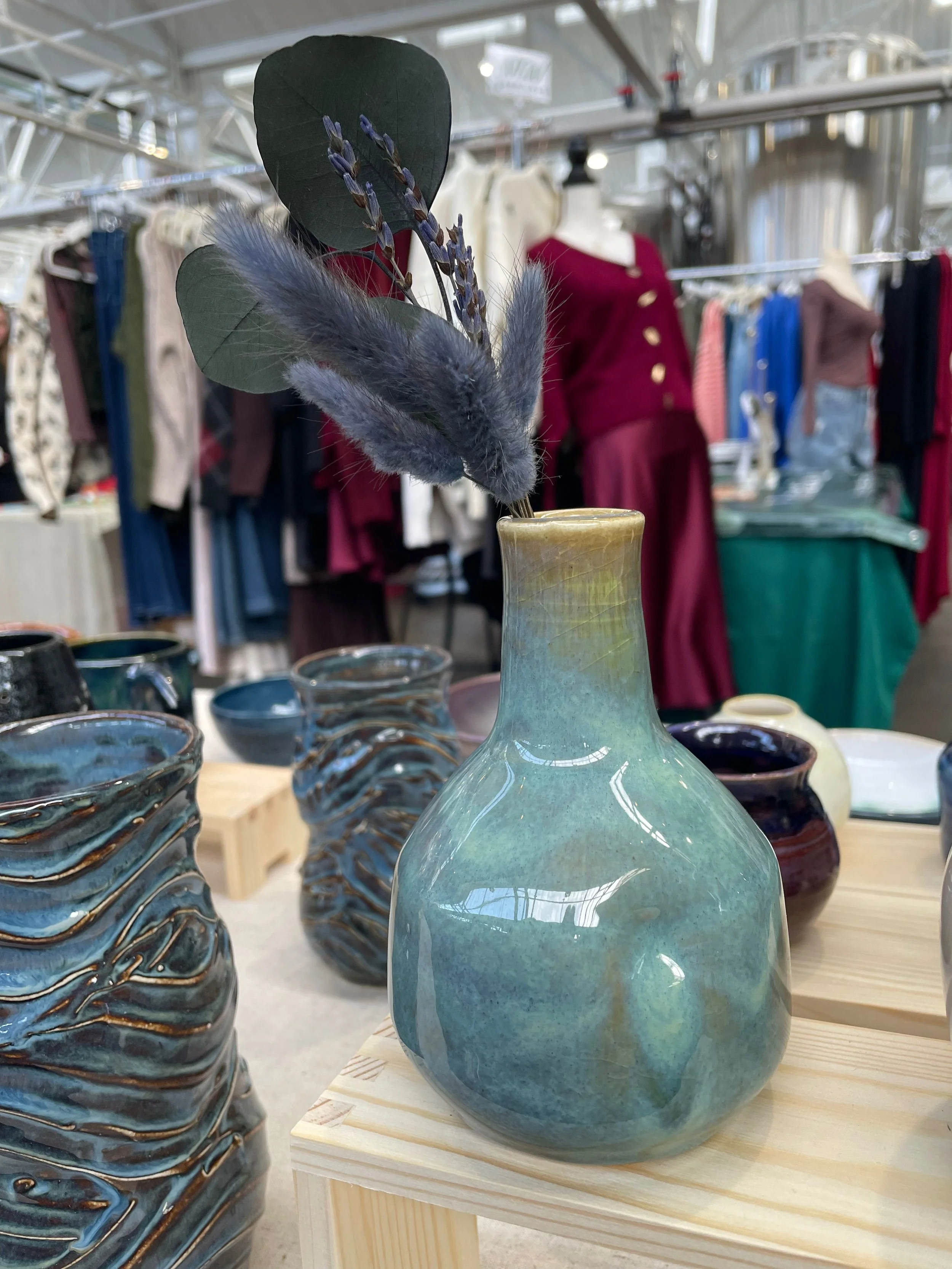 A ceramic vase with a green and blue glaze, filled with dried pampas grass and lavender stems, displayed on a wooden stand at a market.