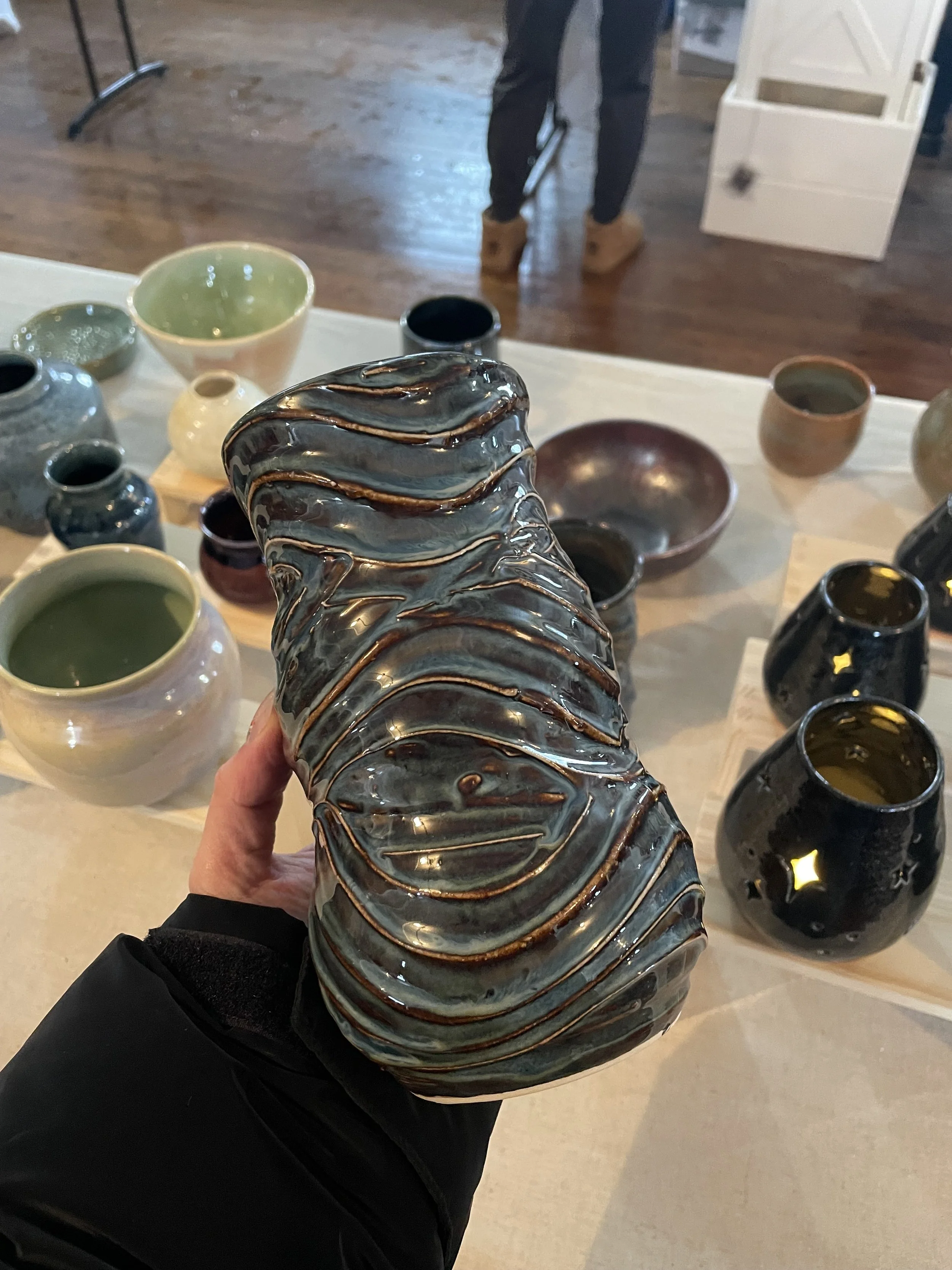 A large, glazed ceramic vase with wavy, textured patterns in front of a display of various ceramic bowls and vases. The table has multiple ceramic items in different colors and shapes.